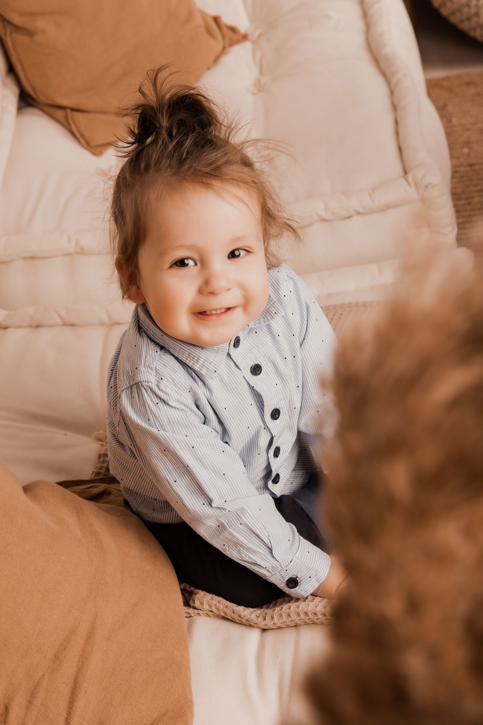 Das Bild zeigt einen kleinen Jungen seitlich von oben fotografiert. Er sitzt auf einem hellen Sofa mit braunen Kissen. Unscharf im Vordergrund ist Pampasgras zu sehen. Der Junge hat braune, lange Haare zu einem Wilden Dutt gebunden und trägt ein hellblaues Hemd mit dunkelblauen Punkten und knöpfen, sowie eine dunkelblaue Hose. Er lächelt und zeigt dabei seine zähne, er hat braune Augen. Er schaut leicht an der Kamera vorbei und ist ca. 1 Jahr alt.