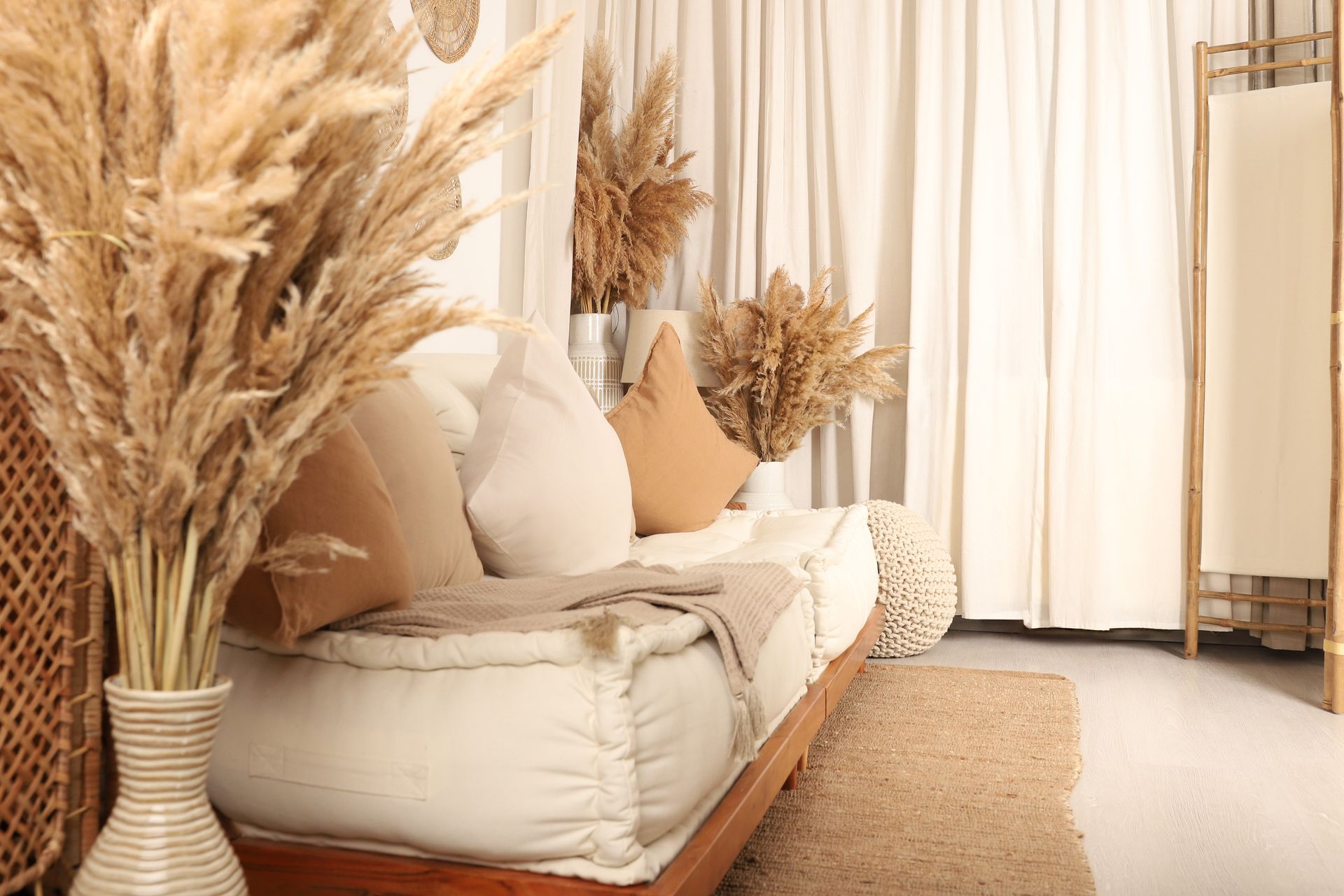 Boho-Stil-Sofa Auf dem Foto ist im schrägen Winkel ein wollweißes Sofa, dekoriert mit beigen und braunen Kissen vor Hellbeigen Vorhängen zu sehen. Im Vorder- und Hintergrund befindet sich Pampasgras als Dekoration.