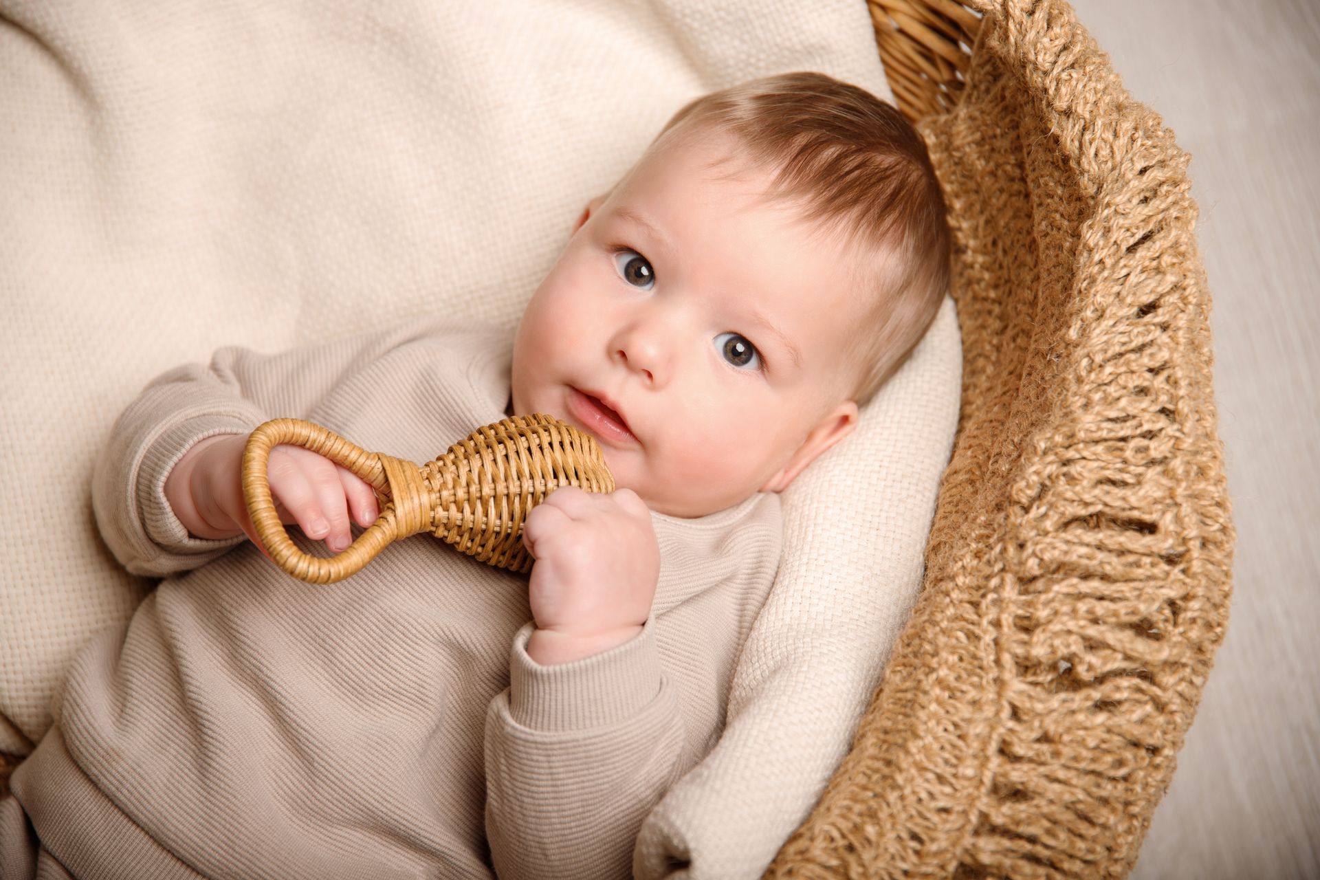 Das Bild zeit ein Baby, Junge, ca. 8 Monate liegend auf einer Beigen Decke von Oben fotografiert. Er hält sich eine Rattanrassel an das Kinn, die er mit beiden hängen festhält und schaut direkt, mit einem freundlichen, interessierten Block in die Kamera. Er hat hellblonde Haare und Braune Augen und liegt auf einer beigen Decke.