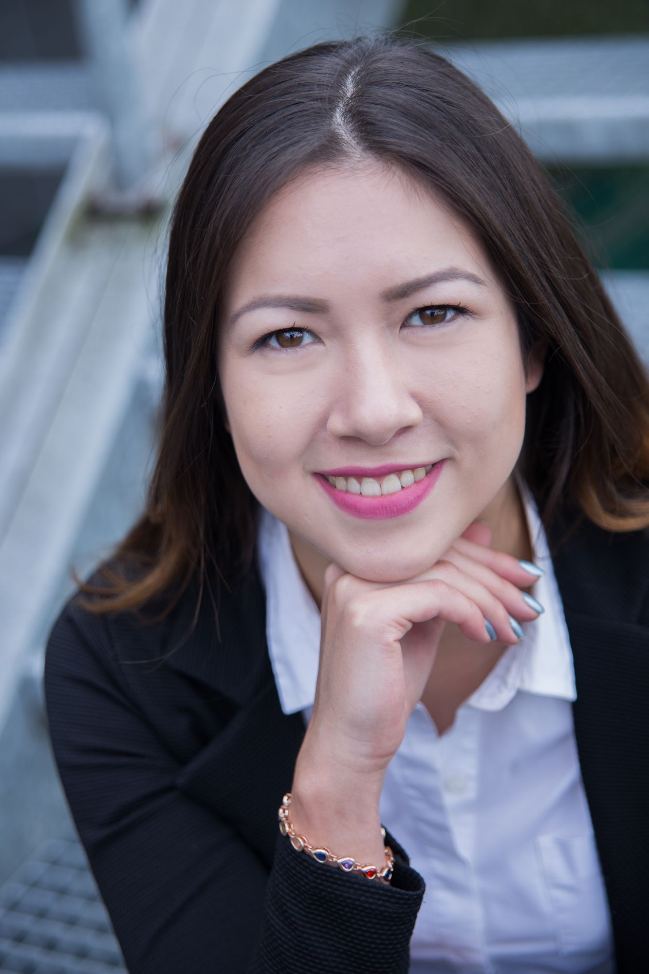 Businessportrait von einer jungen Frau mit dunklen, langen Haaren, die ihr Kinn auf dem Handrücken abstützt. Die Frau trägt pinken Lippenstift, Einen schwarzen Blazer und eine weiße Bluse. Die Farbstimmung ist eher kühl. Die Frau lächelt mit offenem Mund und hat braune Augen.
