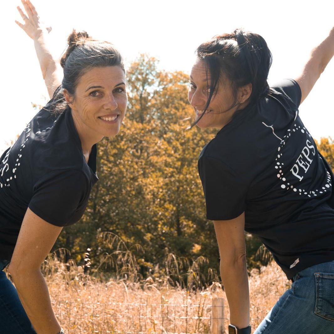 Séverine et Sandrine vêtues de leur tee-shirt PEPS sont souriantes . Elles sont de dos, mais se retournent pour regarder l'objectif avec un bras levé. Photo dynamique.