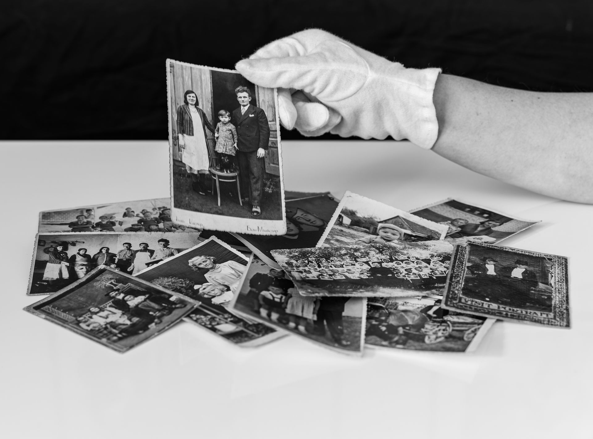 Old photographs are scattered on a table; a gloved hand lifts up one.