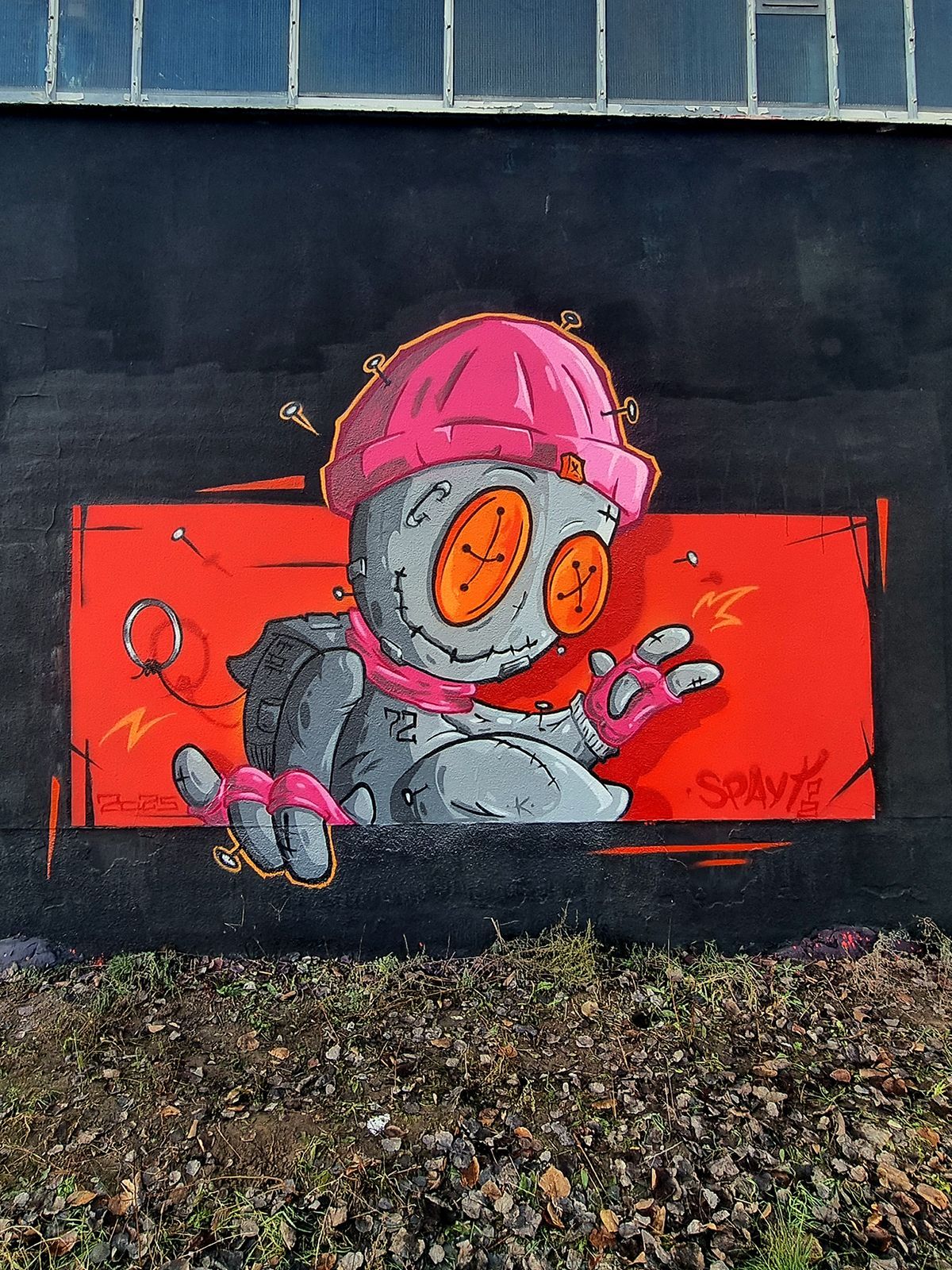 Das Bild zeigt einge gesprühte Voodoopuppe mit Mütze und Rucksack, in Grautönen und mit Akzentfarben vor einem schwarz-orangefarbigem Hintergrund von Spayt72