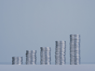 Varios montones de monedas ordenados por cantidades, de izquierda a derecha de menos a más cantidad. Son monedas de plata con un fondo grisáceo con un toque azul.