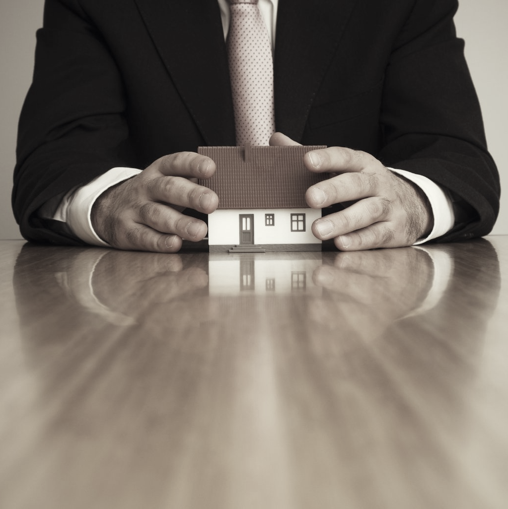 Hombre con traje aguantando una casa miniatura de forma posesiva, como si se la quisiera quedar o apropiar.