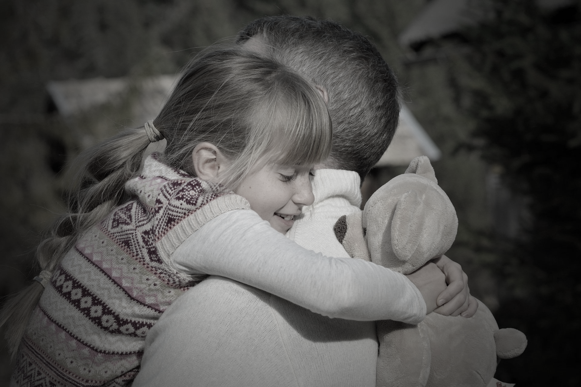 Niña de muy pocos años abrazando a su padre que hace bastante tiempo que no ve a causa del divorcio de sus padres. La niña sonríe mientras abraza a su padre con su osito de peluche.