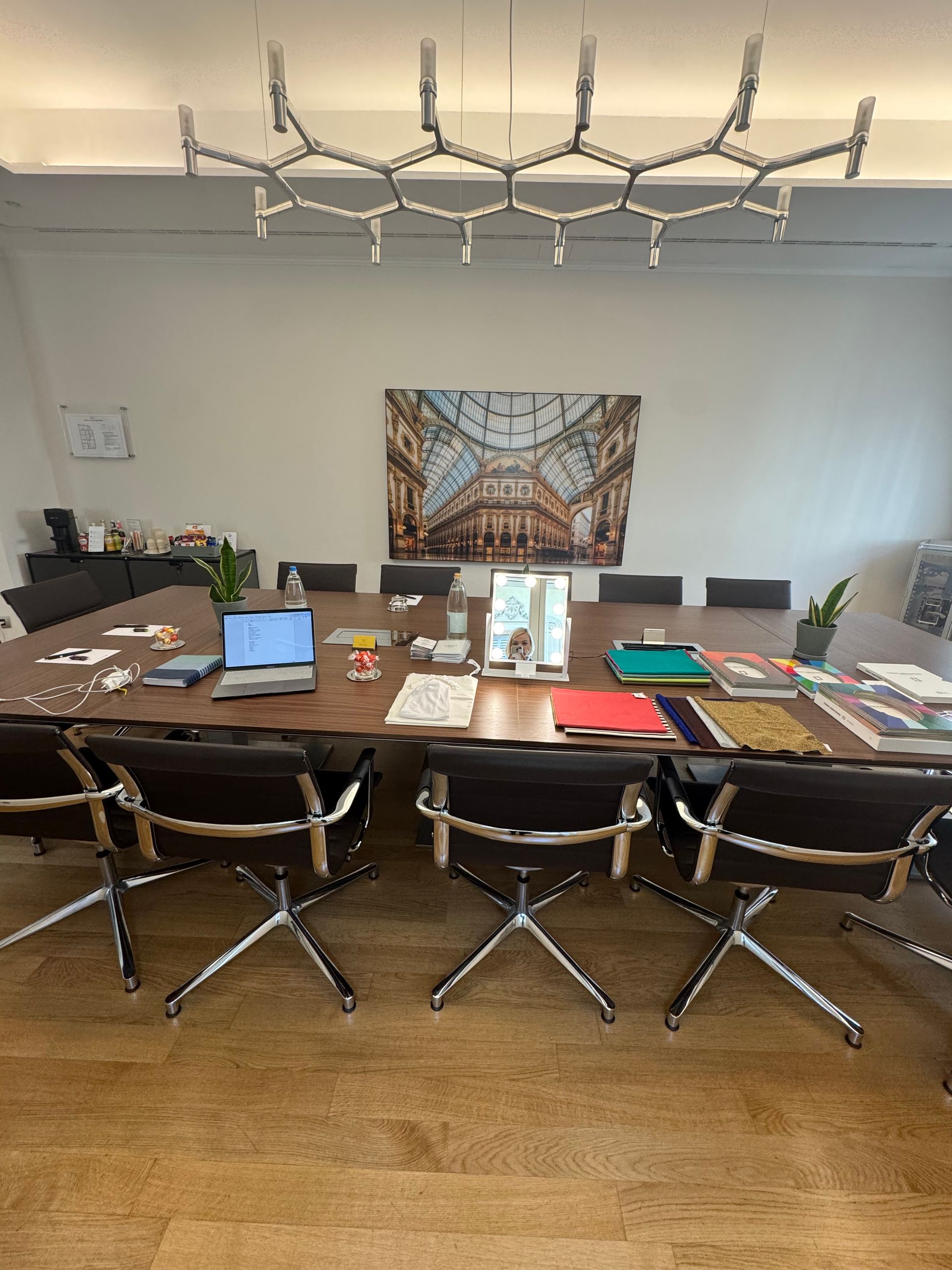 Sala riunioni dell'ufficio in via Montenapoleone, con tavolo rettangolare, sedie, libri, agende, pc e specchio.