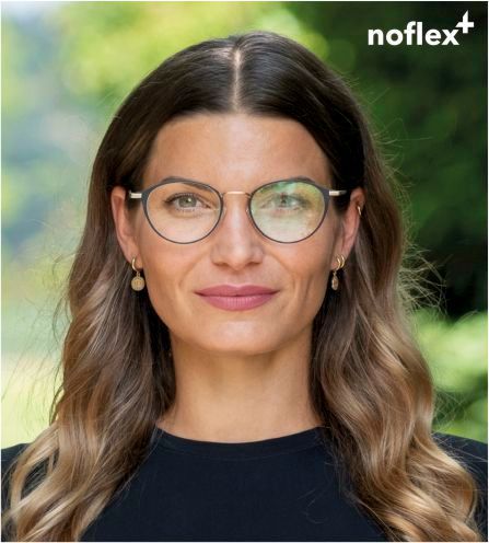 Eine Frau schaut frontal in die Kamera. Sie trägt eine Brille. Das linke Brillenglas ist ohne sichtbaren Reflexe, das rechte Glas dagegen spiegelt die grüne Umgebung ein.