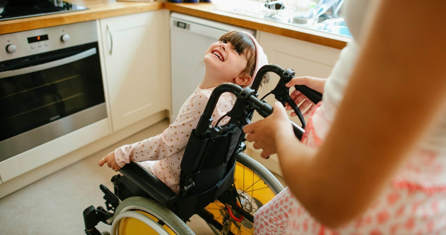 Foto von einem Kind, das in einem Rollstuhl sitzt und die Frau anlächelt, die es schiebt