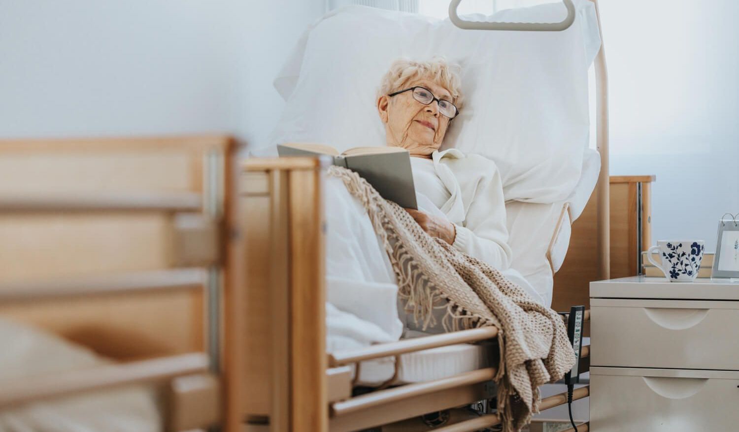 Foto eines Pflegebetts aus Holz, in dem eine ältere Frau liegt, die gerade liest