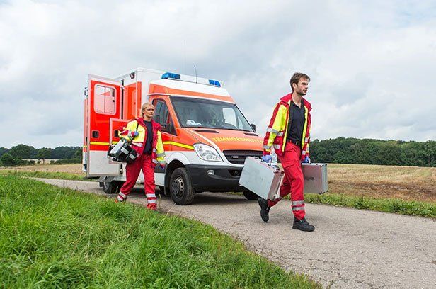 Erste-Hilfe-Kurs für die Berufsausbildung Zwei Sanitäter gehen zum Einsatz. Im Hintergrund steht ein Rettungstransportwagen.