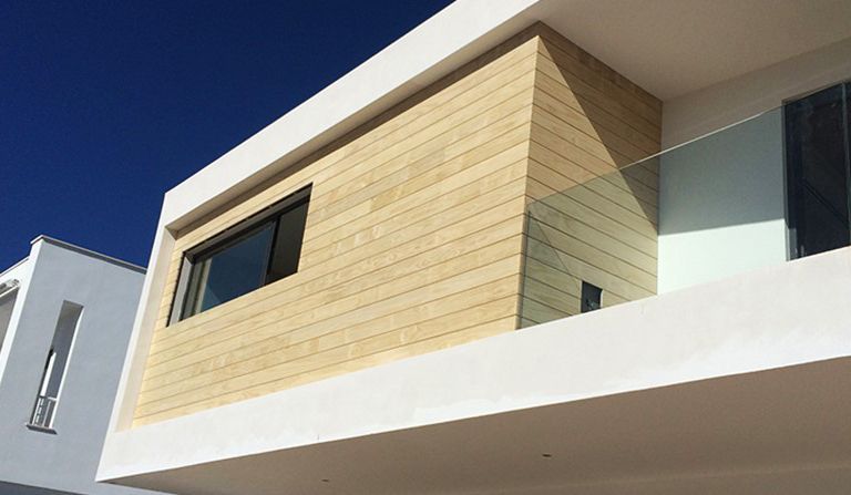 Una casa moderna con un revestimiento de madera exterior de color marrón claro. El revestimiento tiene un acabado texturizado que le da un aspecto natural. La casa tiene un balcón con barandilla de vidrio que ofrece vistas al exterior.