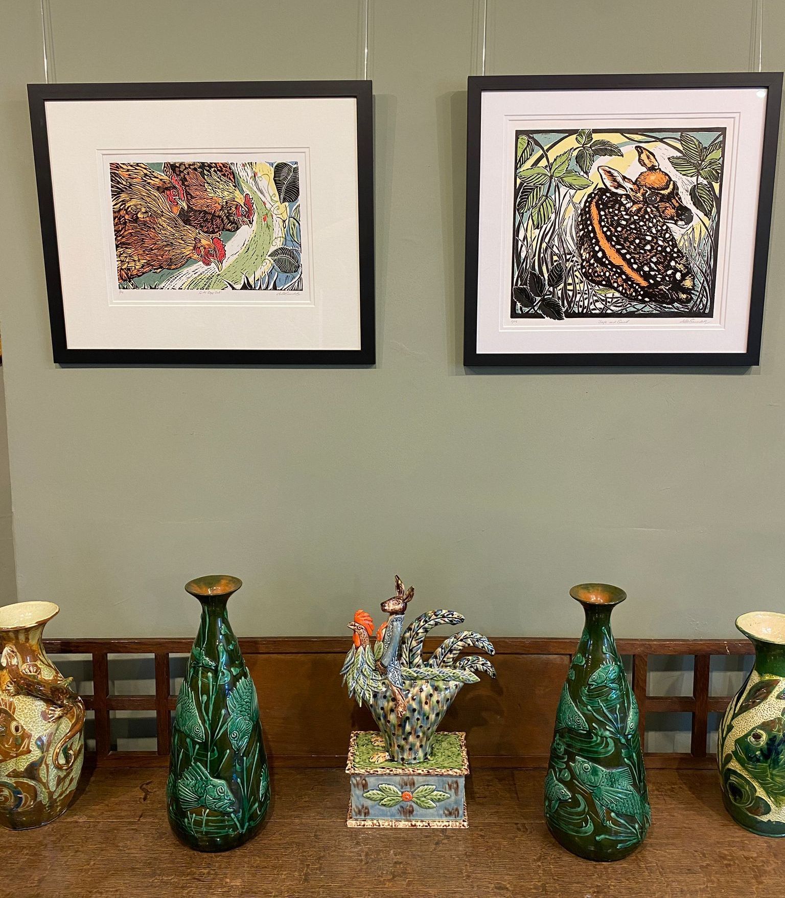 A photograph of two framed colourful lino prints. The one on the left is of three chickens, the one on the right is of a deer fawn. The pictures are hanging on a sage green painted wall. They are hanging above a wooden cabinet with five ceramic pieces on display. There are four vases - two brown and cream and two jade green patterned pieces. A ceramic hare riding a brightly glazed cockerel sits in the middle, between the two green vases.