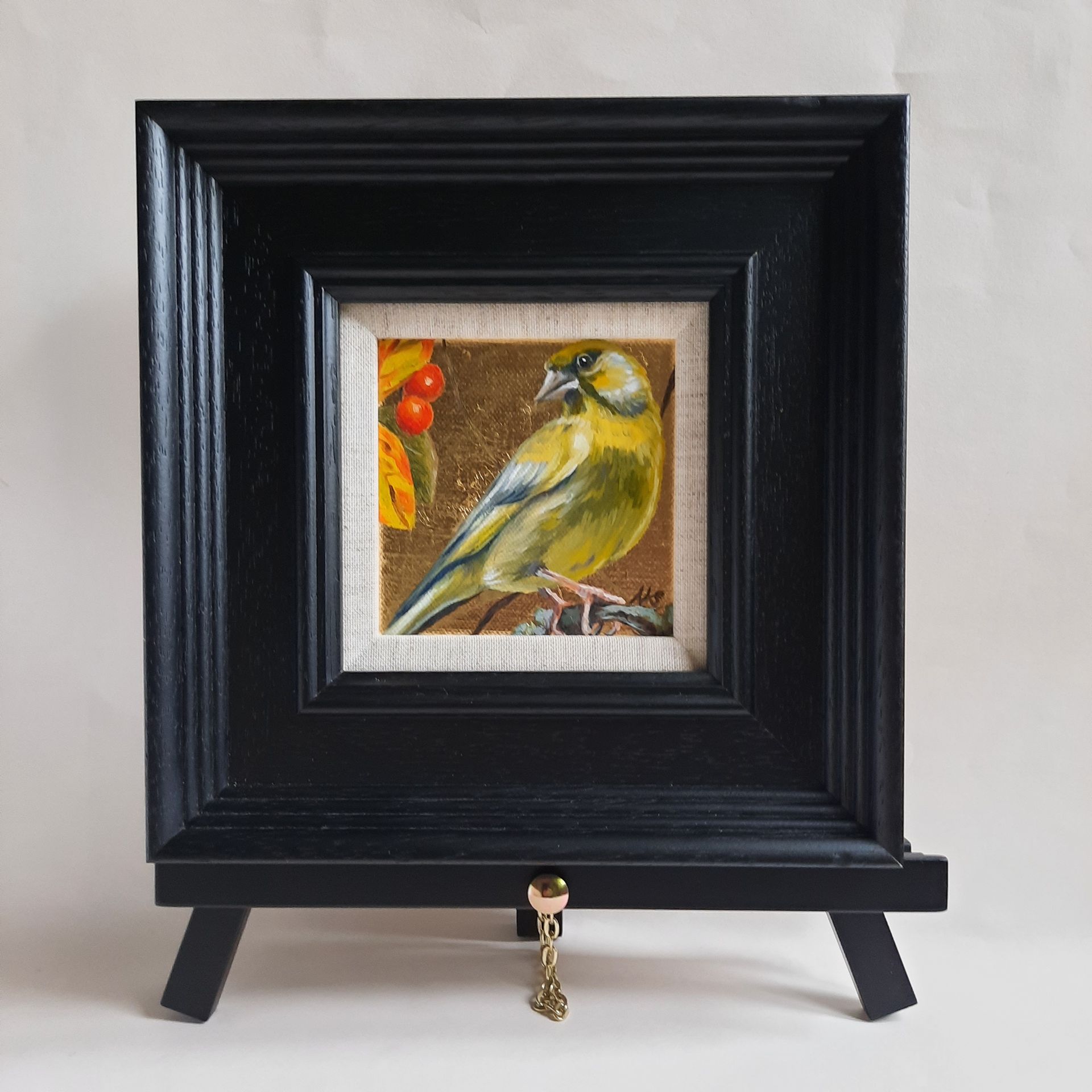 An oil painting of a Green Finch perched on a twig, with round, bright orange berries and autumnal leaves in the top left corner. The painting is on shiny gold leaf and is shown framed in a black frame on a black mini easel