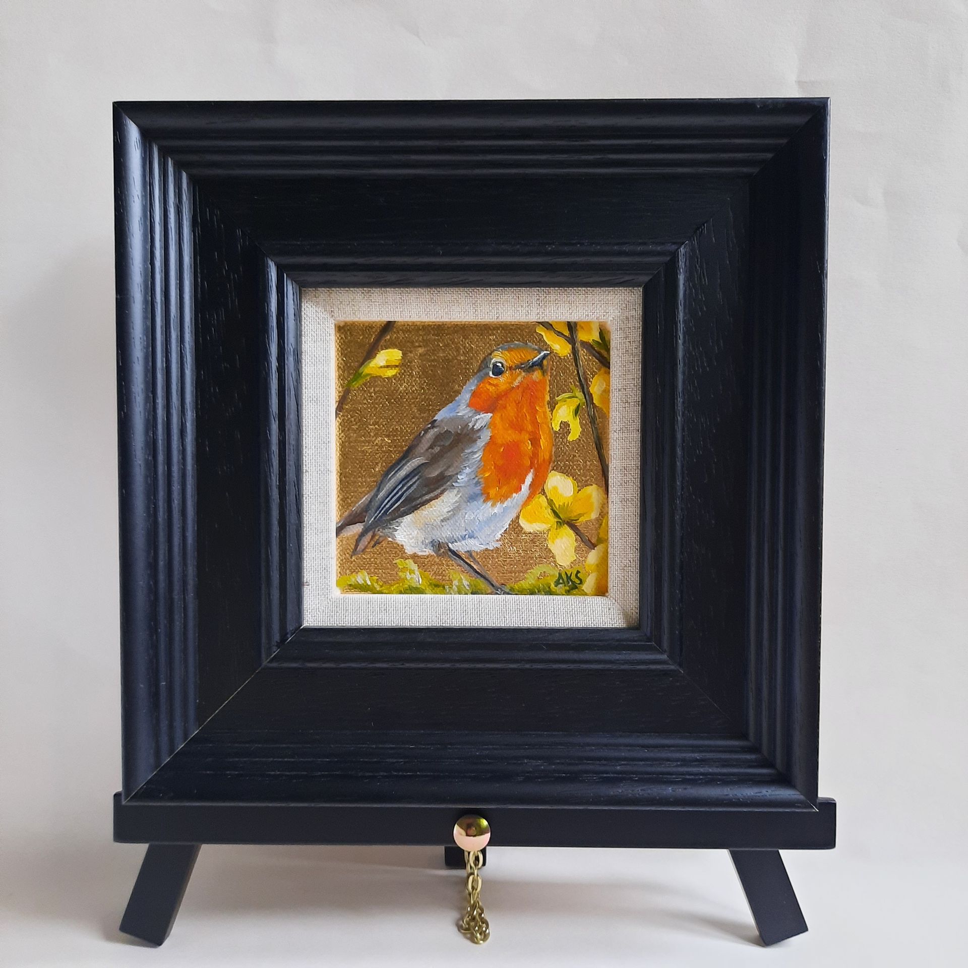 An oil painting of a red breasted Robin bird perched on a twig, surrounded by bright yellow Forsythia flowers. The painting is on shiny gold leaf and is shown framed in a black frame on a black mini easel