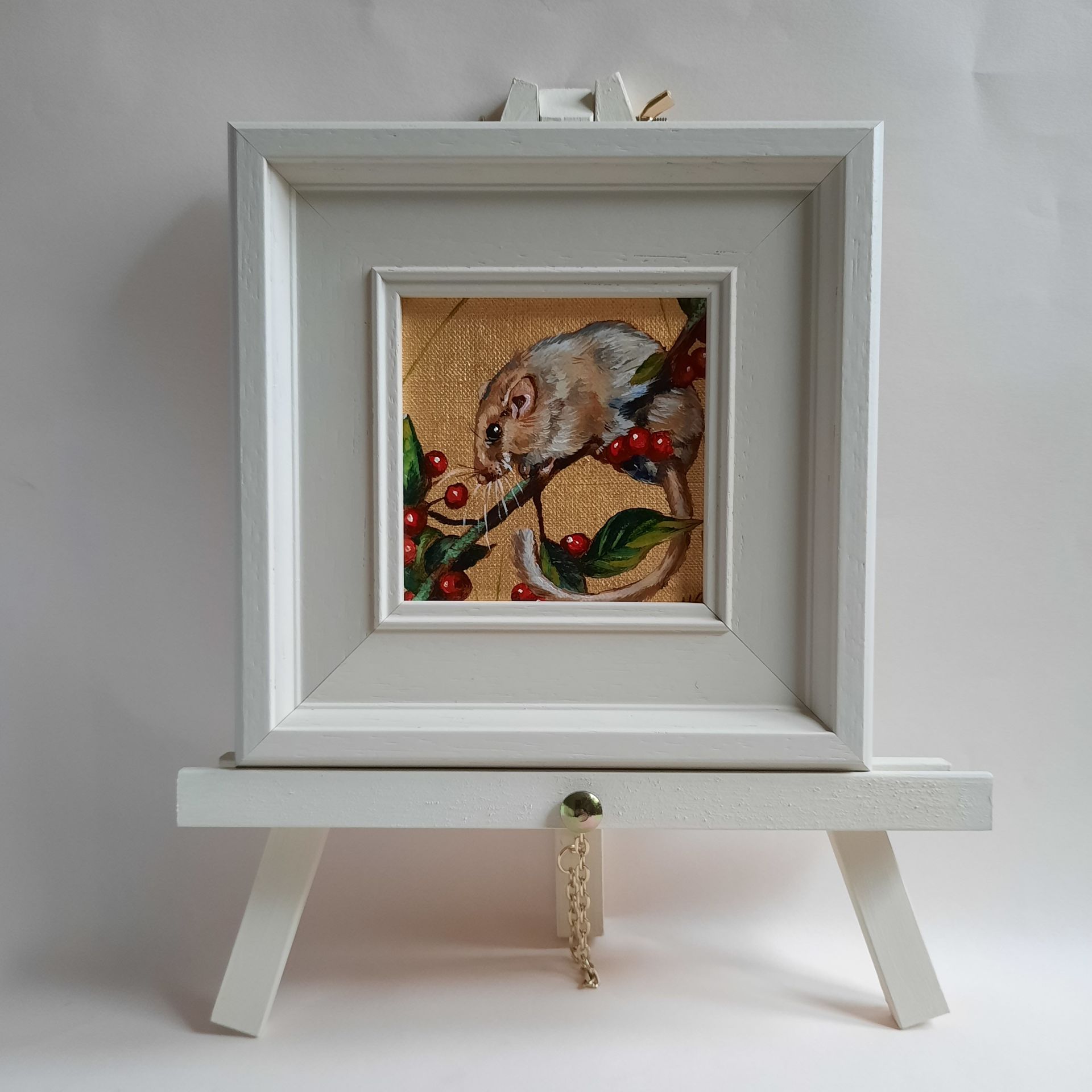 An oil painting of a Dormouse in profile, balanced on a twig. Dark green leaves and bright red, round berries surround it. The painting is on shiny gold leaf and is shown framed in a cream coloured frame on a matching mini easel