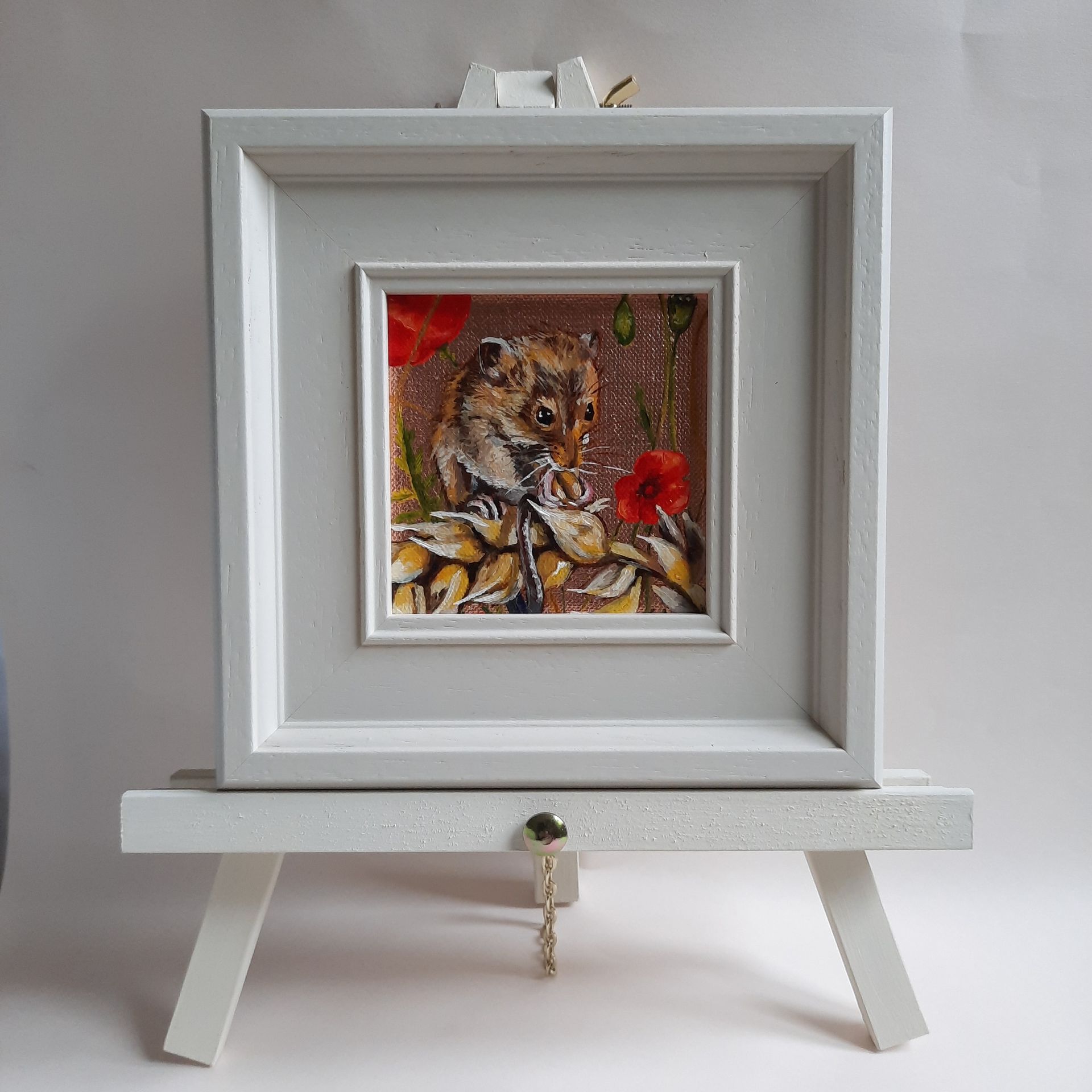 An oil painting of a Harvest Mouse sitting on a curved ear of wheat, eating a grain. Bright red poppy flowers are in the background. The painting is on shiny copper leaf and is shown framed in a cream coloured frame on a matching mini easel