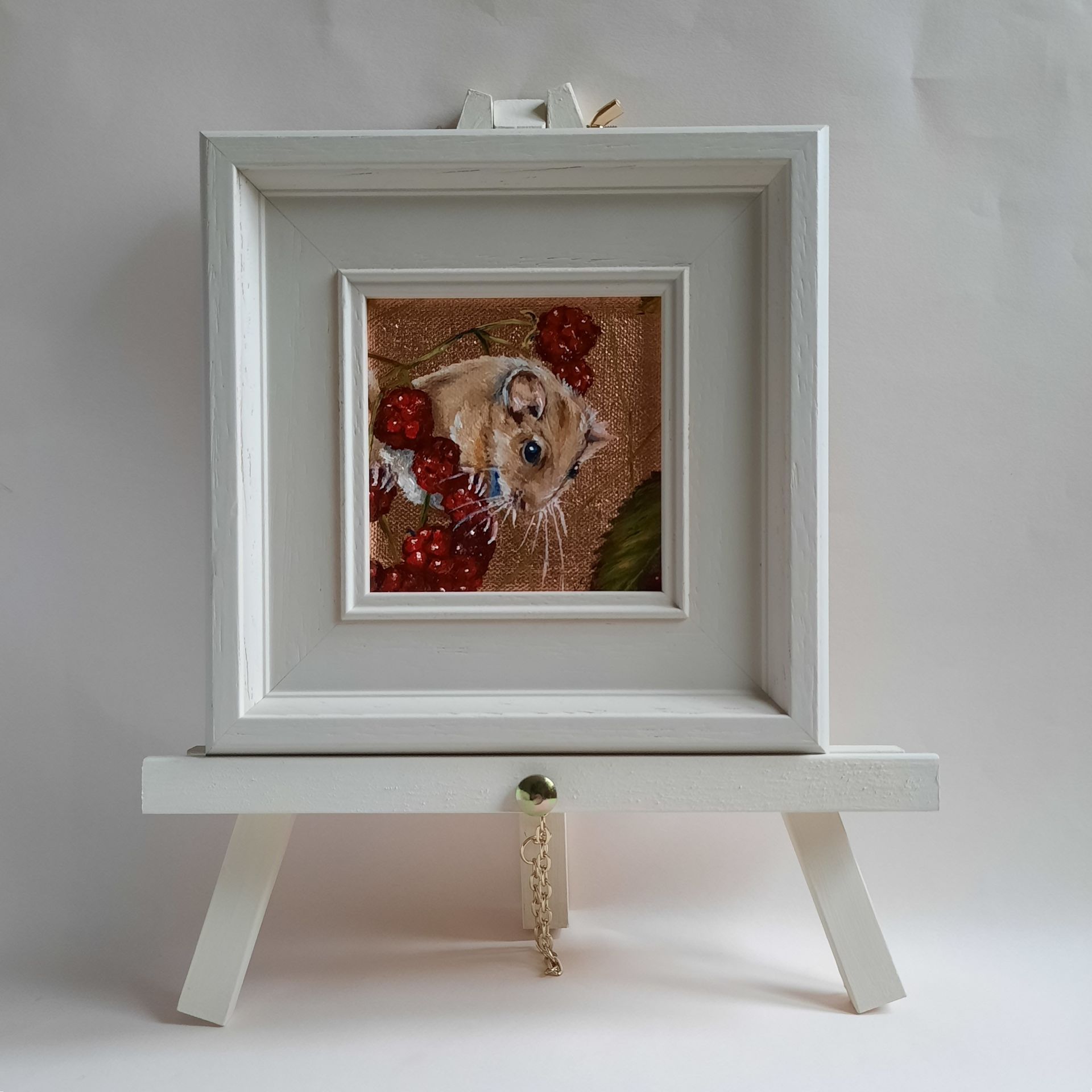 An oil painting of a plump Dormouse holding on to a sprig of red brambles. The edge of a green leaf can be made out in the bottom right corner. The painting is on shiny copper leaf and is shown framed in a cream coloured frame on a matching mini easel