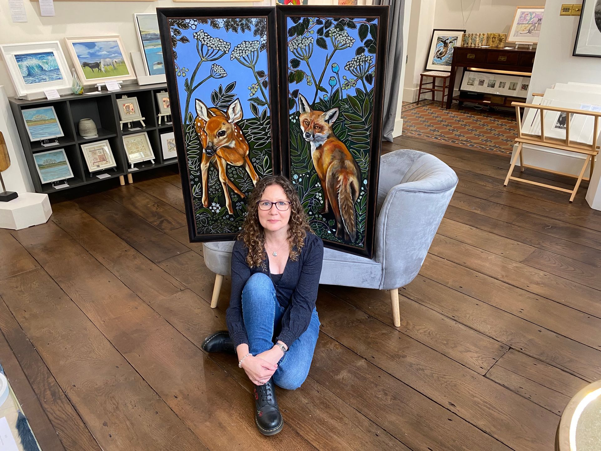 A photo of artist Anita Saunders sitting on the wooden floor of an art gallery in front of two of her paintings. A young deer is the subject of the painting on the left and a fox, the subject of the painting on the right.