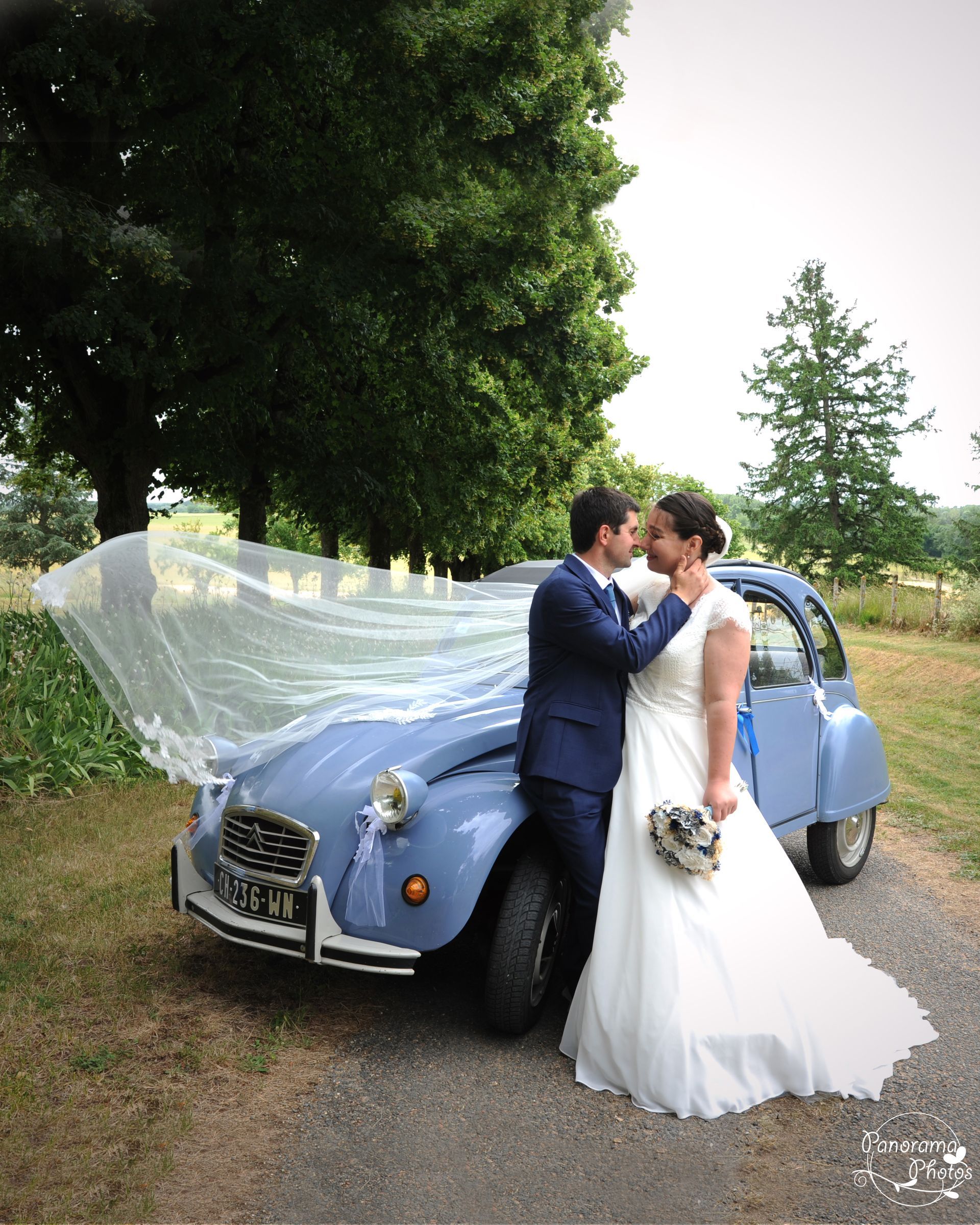 couple de marié devant une voiture 2 CV. Le voile de la marié s'envole avec le vent