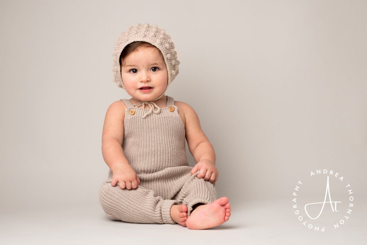 A sitting baby wearing a timless beige wool dungaree set and bonnet during a sitter portrait session