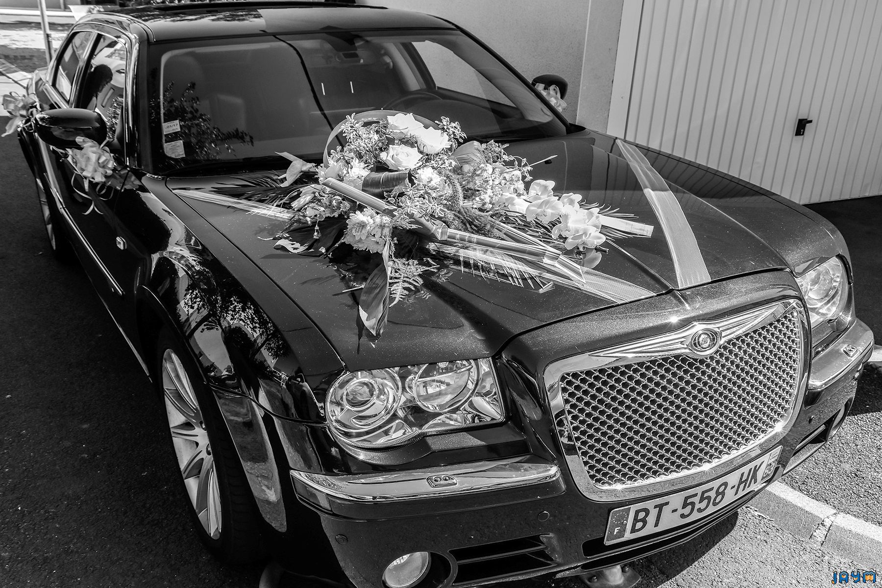 Image en noir et blanc du cortège de mariage : la Chrysler décorée et les rubans de voiture, avec la plaque d'immatriculation française visible.