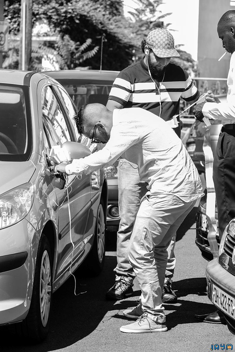 Photo de reportage de mariage en noir et blanc : amis préparant et décorant la voiture verte du cortège avec des rubans en extérieur.