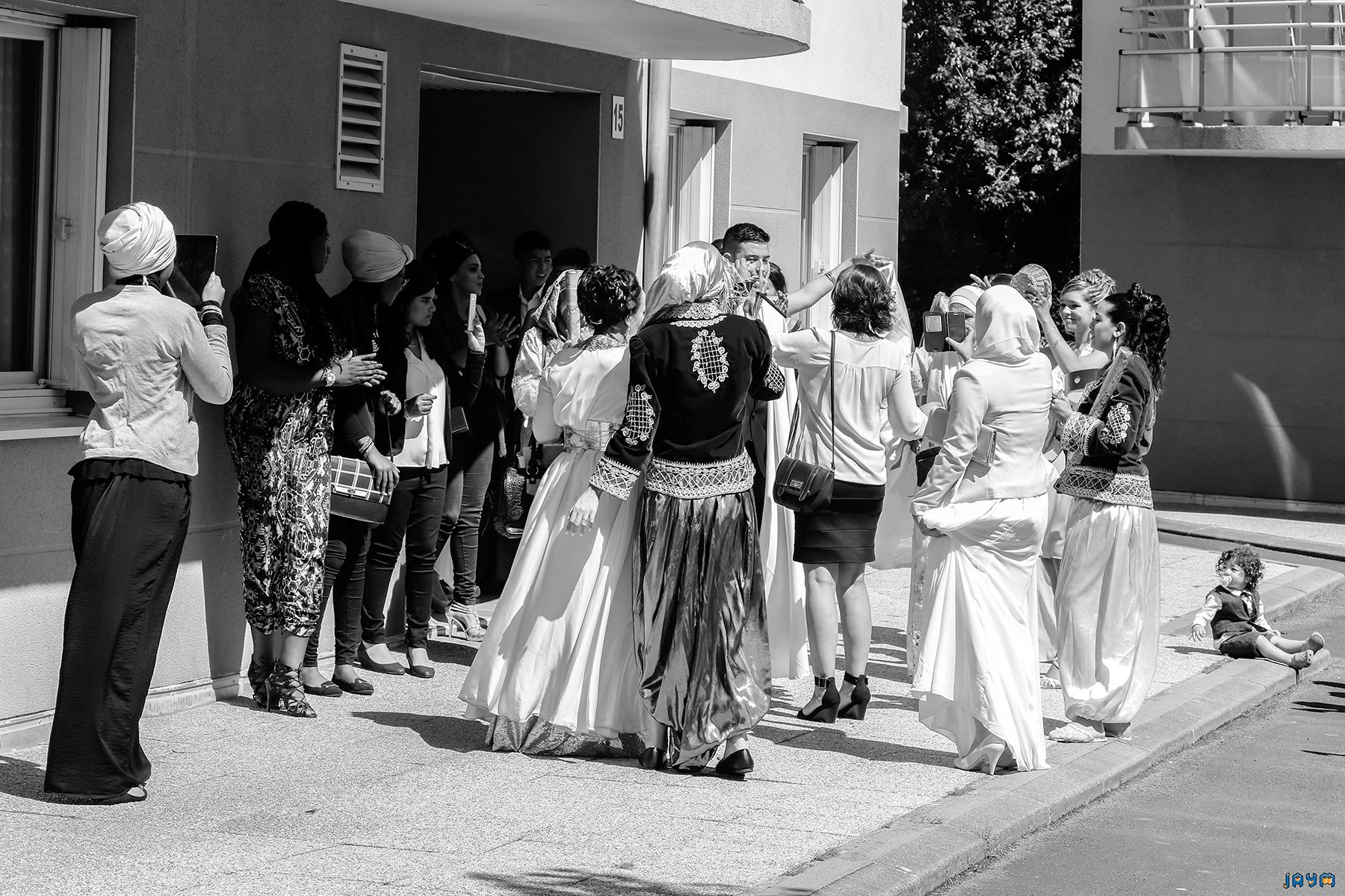 Image en noir et blanc d'un rassemblement de mariage : la mariée en robe blanche et le marié en costume, souriant au milieu des invités.