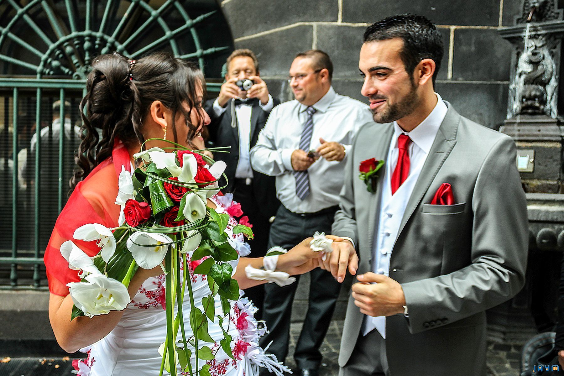 Clémentine et Nicolas, les mariés du jour, échangent un regard rempli d'amour et de complicité. Cette photo de mariage en couleur, prise devant la fontaine historique de Clermont-Ferrand, capture l'émotion de leur union. Le costume de marié gris avec sa cravate rouge et le bouquet de la mariée apportent une touche de couleur vibrante à cet instant inoubliable de leur cérémonie de célébration.