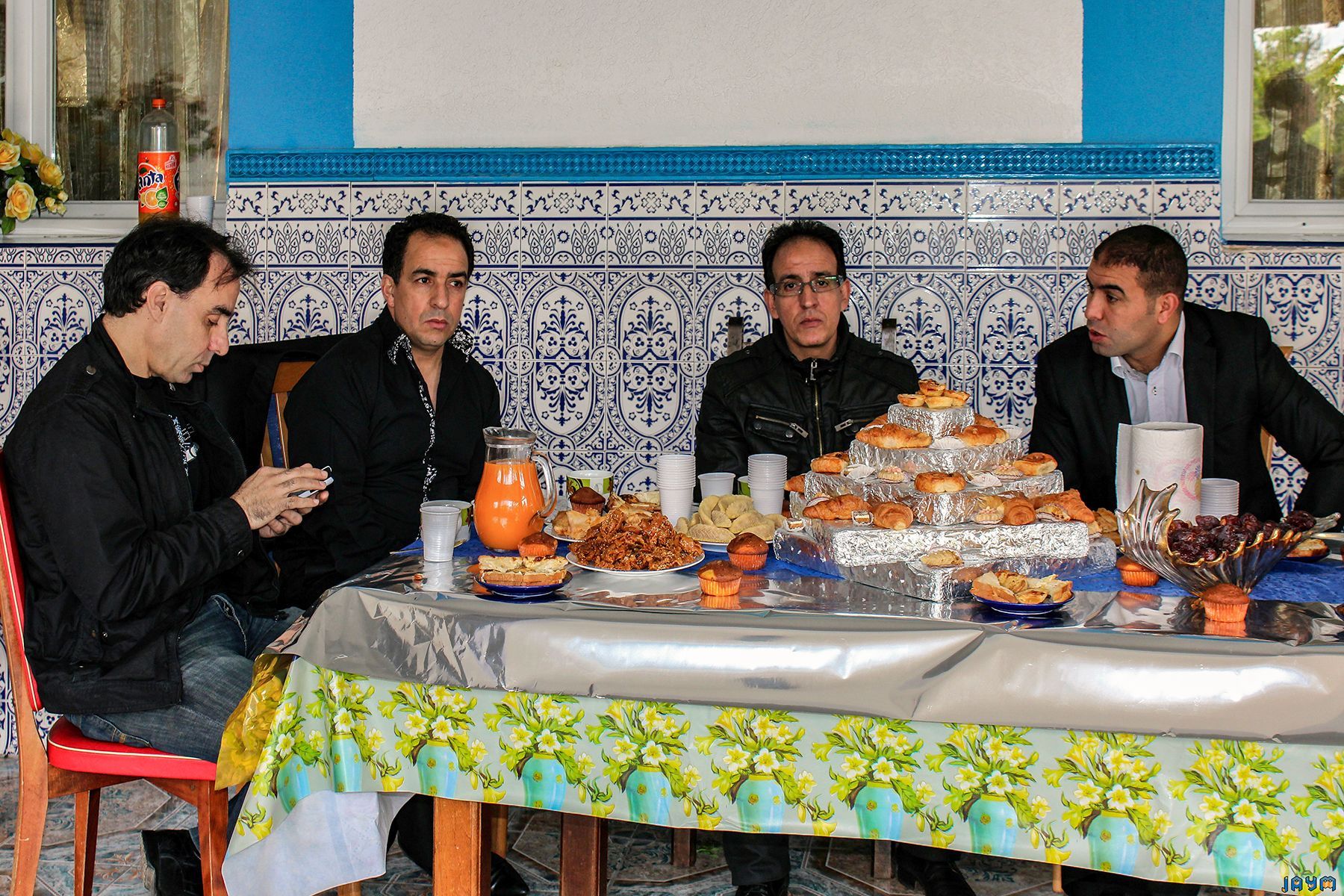 Quatre hommes attablés autour d'un grand buffet composé de pâtisseries, jus d'orange et dattes, devant un mur décoré de carrelage bleu et blanc.
