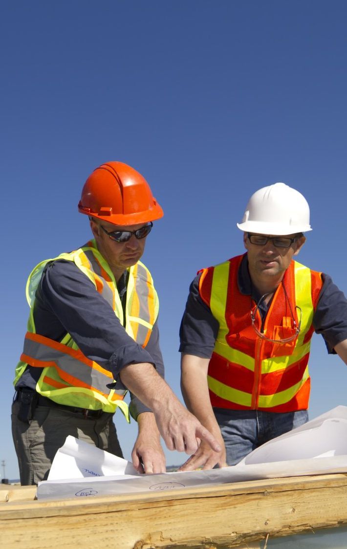Two construction workers wearing safety vests and hard hats reviewing blueprints on a construction site under a clear blue sky.