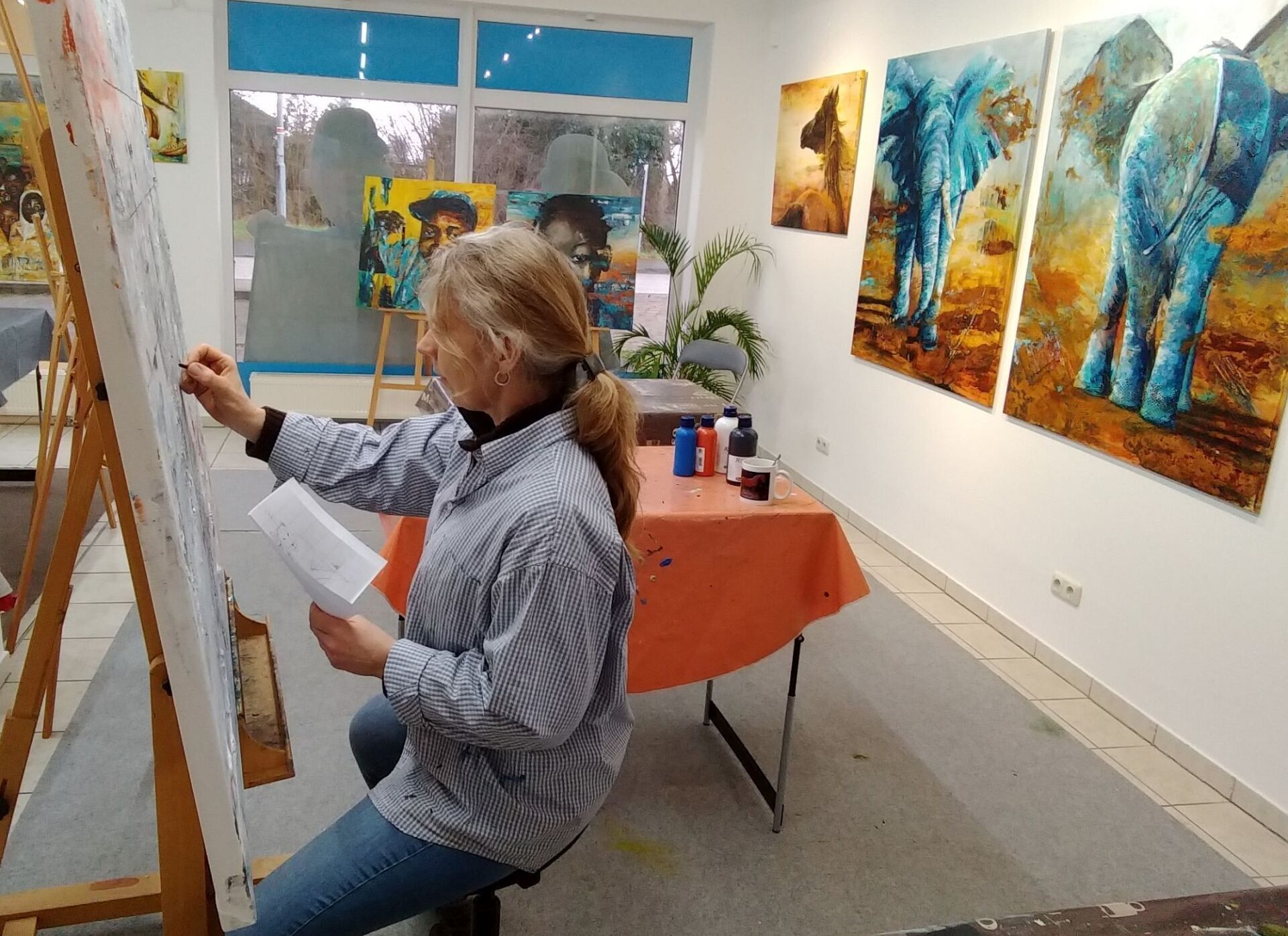 Bianca Leidner an der Staffelei bei einer Auftragsarbeit in ihrem Atelier. Im Hintergrund Bilder mit Elefanten