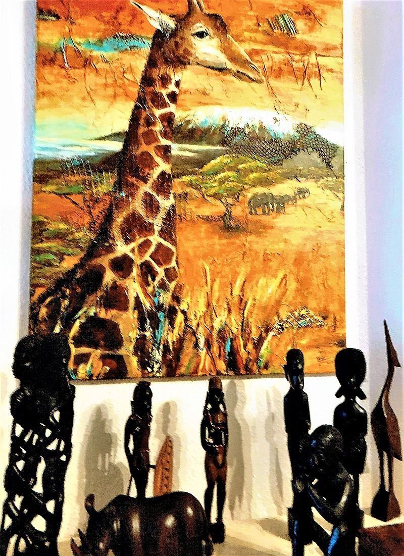 Gemälde in warmen Farbtönen orange, gelb, grün mit einer Giraffe im Vordergrund. Im Hintergrund eine Herde Elefanten und der Kilimandscharo. Vor dem Bild stehen afrikanische Holzfiguren.
