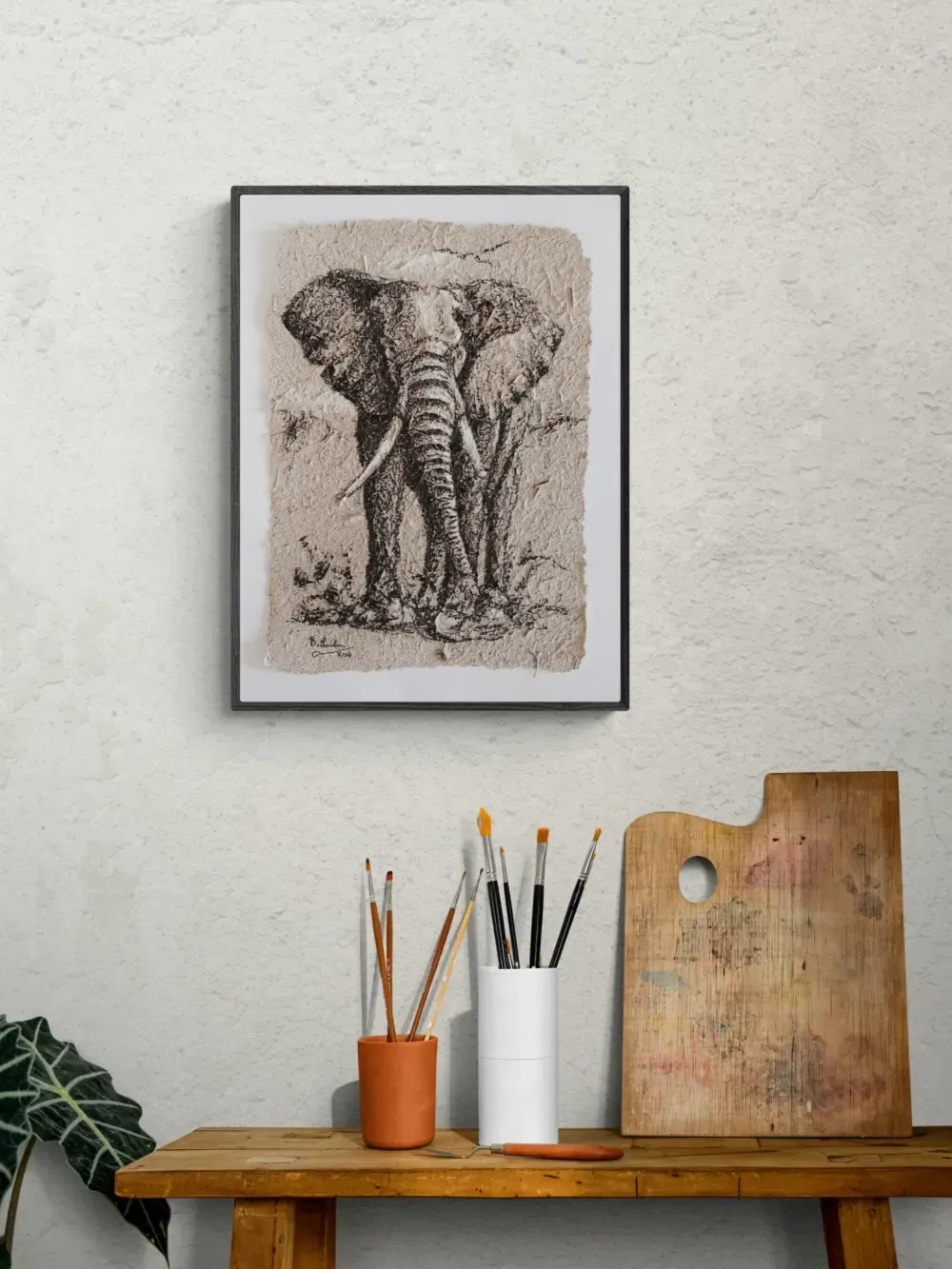 Gerahmte Kohlezeichnug eines Elefanten auf Elefantenpapier aus Elefantendung. Es hängt an der Wand über einem Tisch mit Malutensilien darauf.