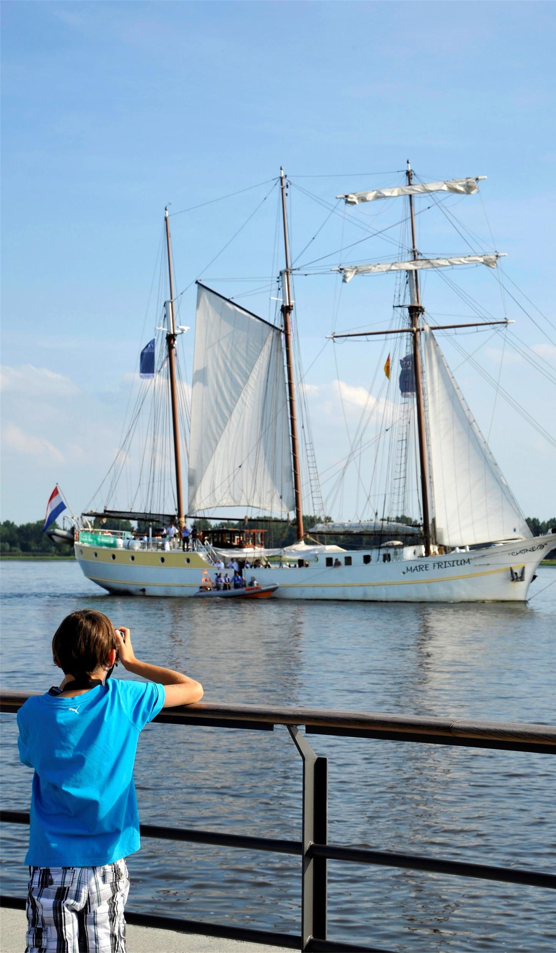 Auf dem Bild ist im Vordergrund ein Junge von hinten zu sehen, deraufs Wasser blickt. Im Hintergrund fährt auf dem Wasser ein dreimastiges altes Schiff vorbei.