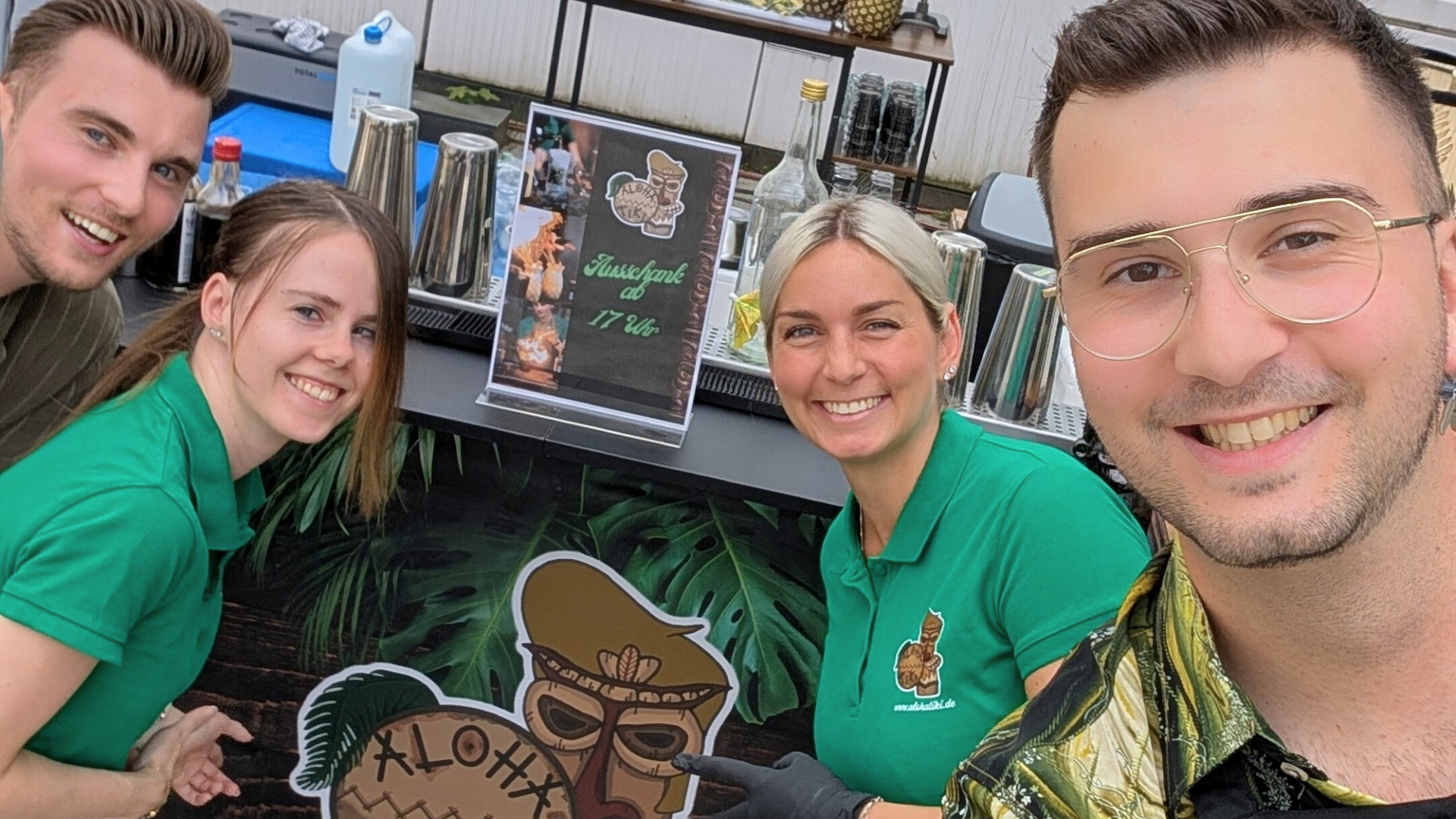 Das ALOHATIKI Team mit mobiler Cocktailbar auf einem Event, lachende Barkeeper in grünen Poloshirts