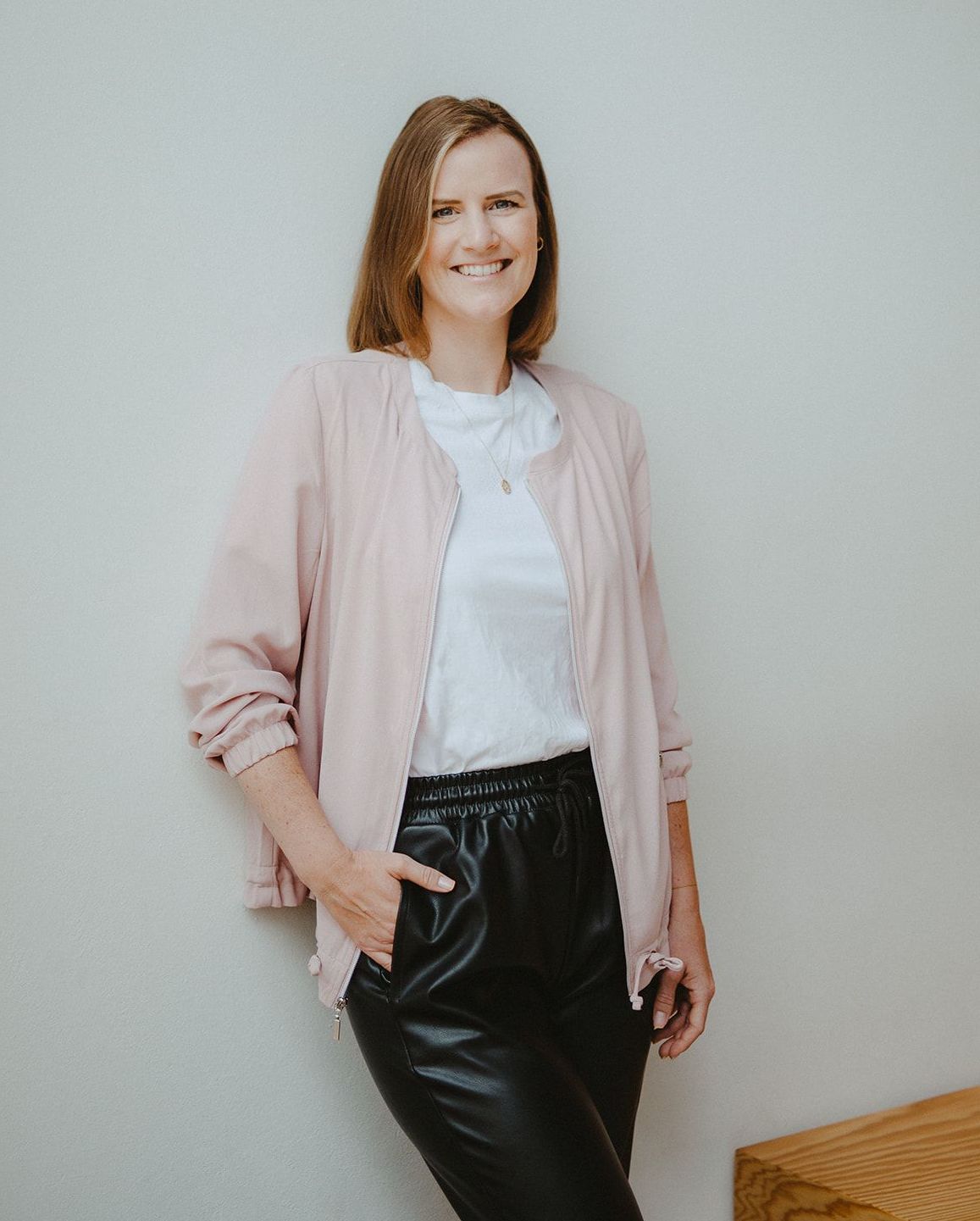 Anna, Texterin und SEO Coach Brünette Frau mit weißem T-Shirt, rosa Jacke und schwarzer Hose lehnt an der Wand und lächelt in die Kamera.