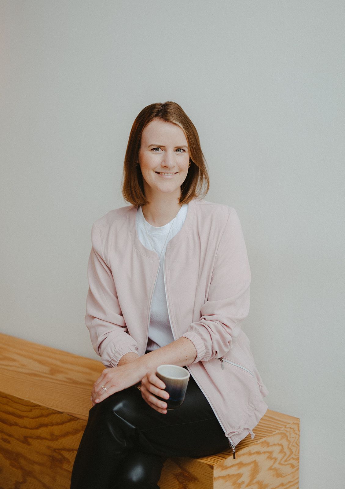Ich liebe Kaffee, aber bitte mit Kokosmilch! Brünette Frau mit rosa Jacke und weißem T-Shirt sitzt auf Holzbank und hält Kaffeebecher in der Hand. Sie lächelt in die Kamera.