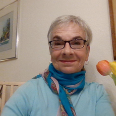 Photograph of Mendy McLean Stone. A smiling silver haired woman wearing dark rimmed glasses, a dark turquoise scarf and a light turquoise top.
