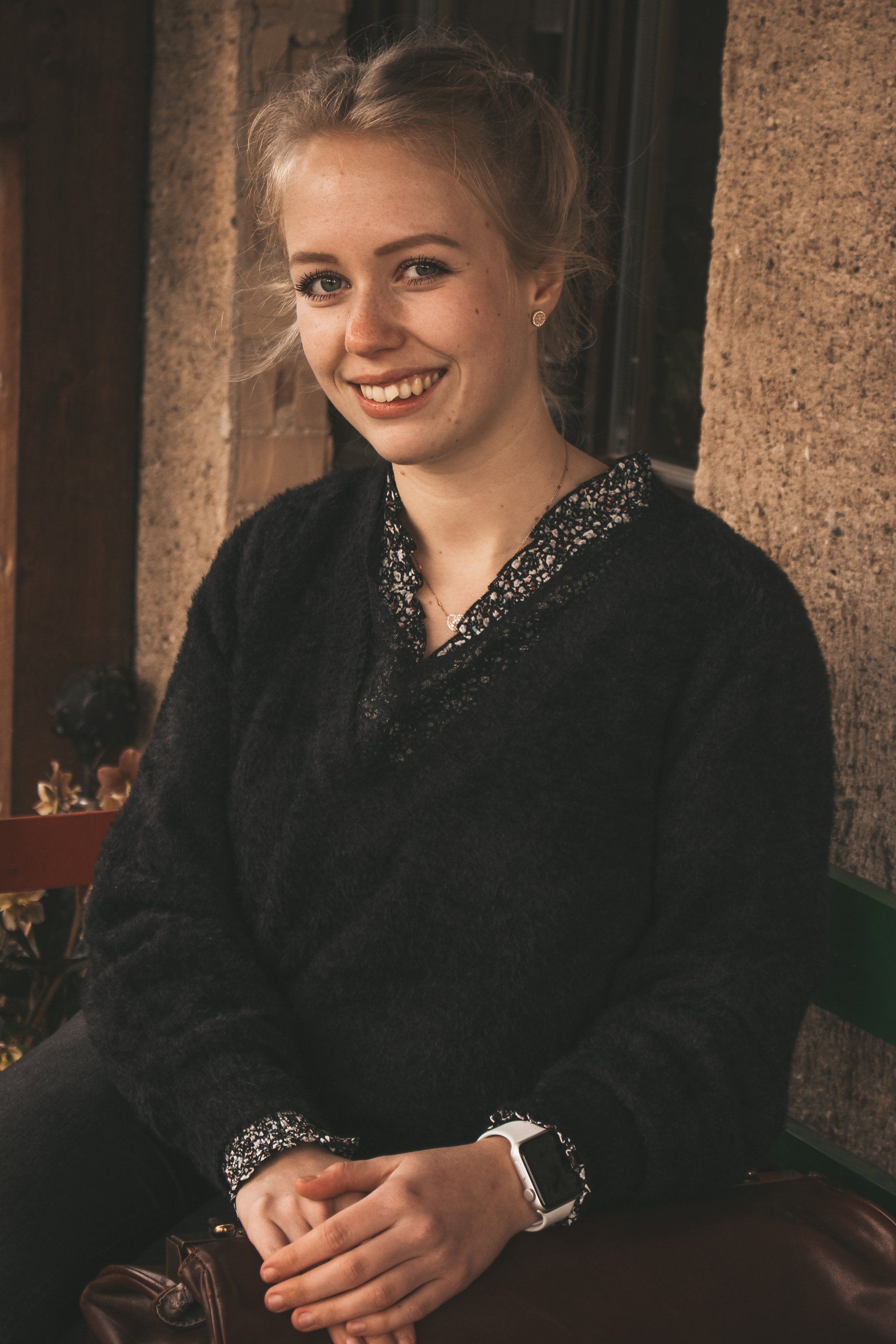 Hebamme Sophie sitzt auf einer Bank vor der Hauswand und lächelt in die Kamera. Links von ihr steht ihr Hebammenkoffer, auf dem ihre Hände liegen.