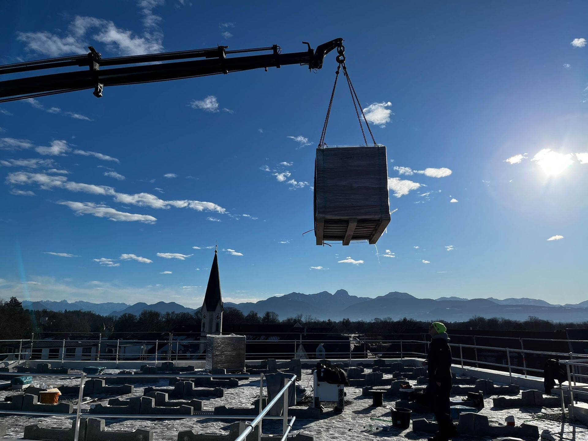 Kran hebt verpackte Palette auf verschneites Dach, Arbeiter beobachtet, Alpen und Kirchturm im Hinte