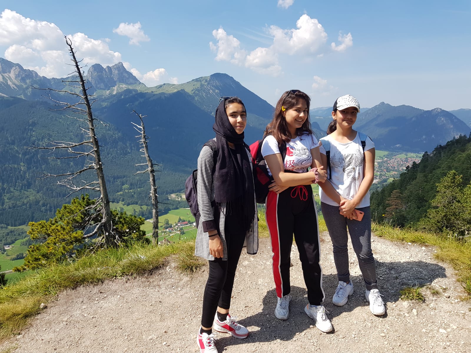 Drei Mädchen stehen auf einem Berg. Hinter ihnen sieht man das Alpenpanorama. Die Sonne scheint.