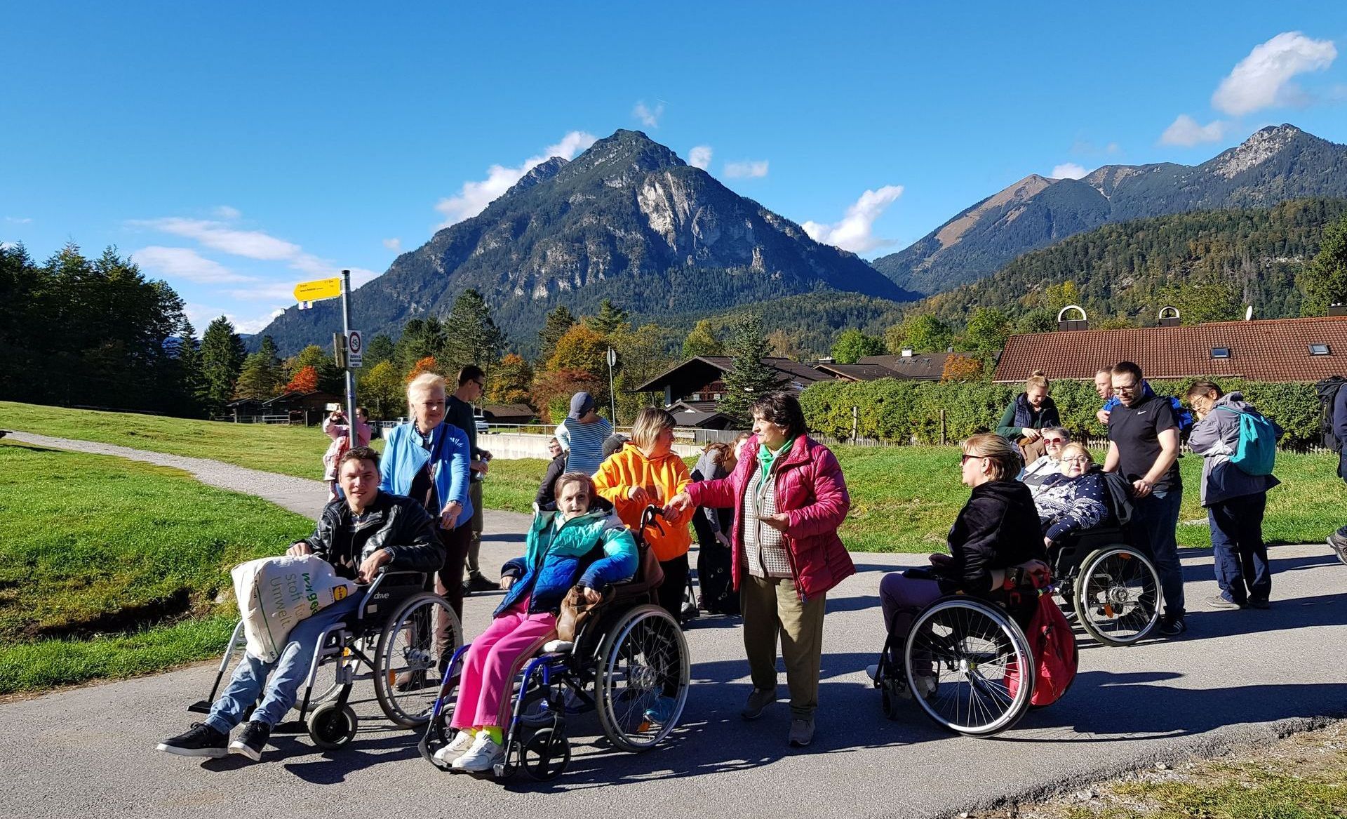 Eine Gruppe von Menschen ist im Bild zu sehen. Einige nutzen einen Rollstuhl. Sie gehen auf einer Straße. Im Hintergrund ist eine Bergkette zu sehen.