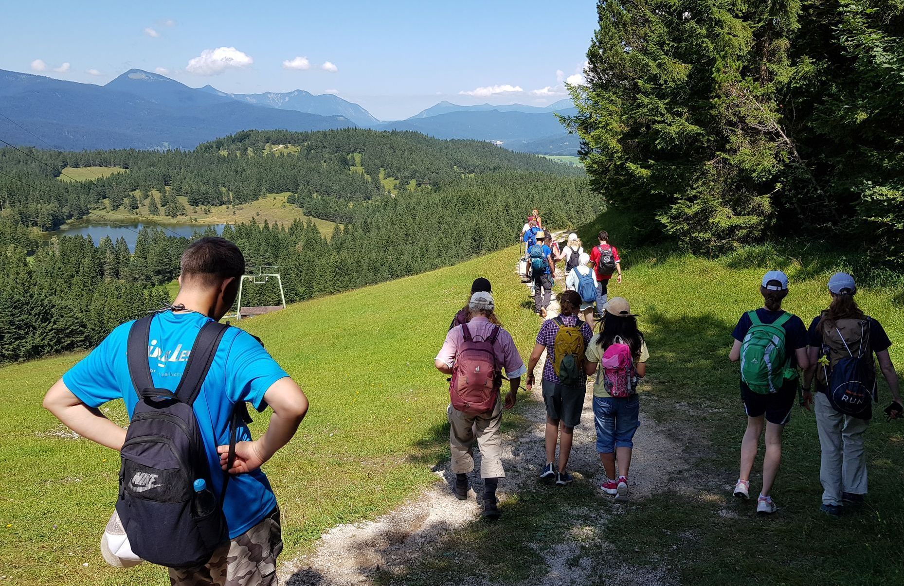 Es ist eine Wandergruppe von hinten zu sehen. Im Hintergrund sieht man am Horizont eine Bergkette. Im Vordergrund ist eine grüne Wiesenfläche und es sind Bäume zu sehen.