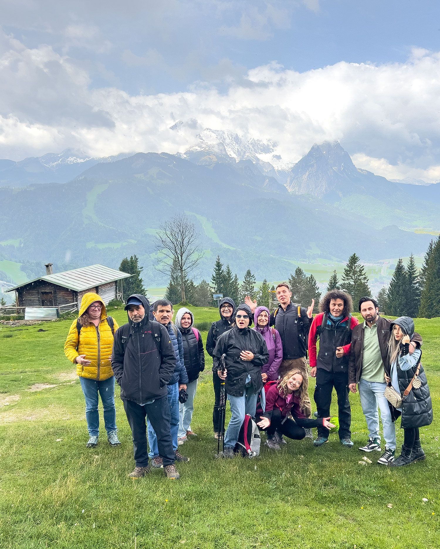 Gruppe von 12 Personen im Freien, vor einer Berglandschaft mit schneebedeckten Gipfeln. Im Hintergrund eine Holzstruktur. Einige tragen Jacken in verschiedenen Farben.