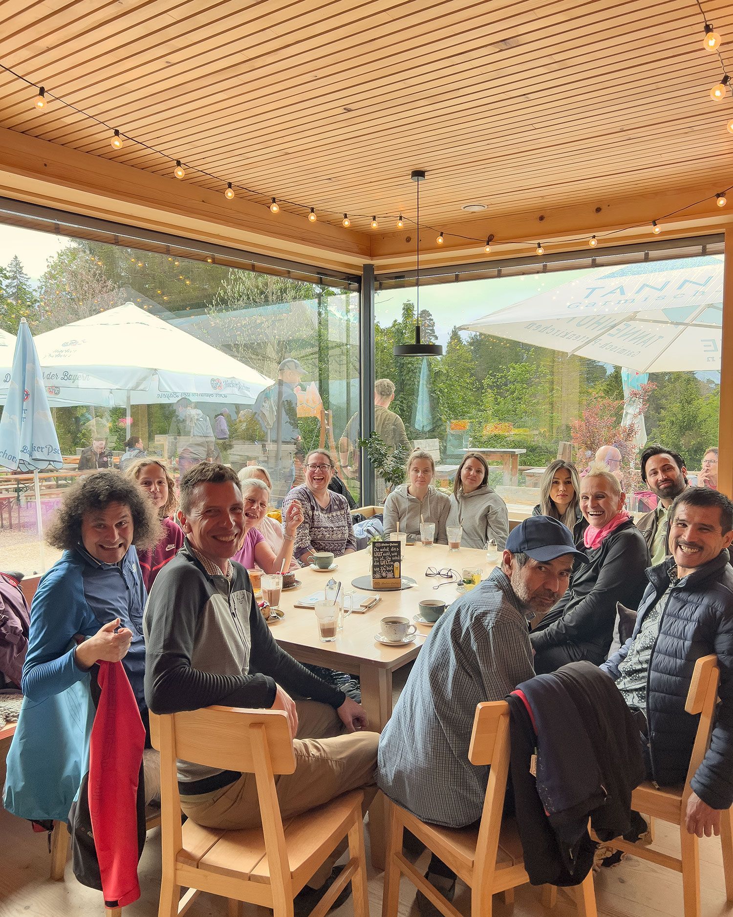 Eine Gruppe von 14 Personen sitzt an einem runden Tisch in einem hellen Raum mit Holzdecke. Auf dem Tisch stehen Getränke und Geschirr. Im Hintergrund sind Sonnenschirme und Bäume sichtbar.