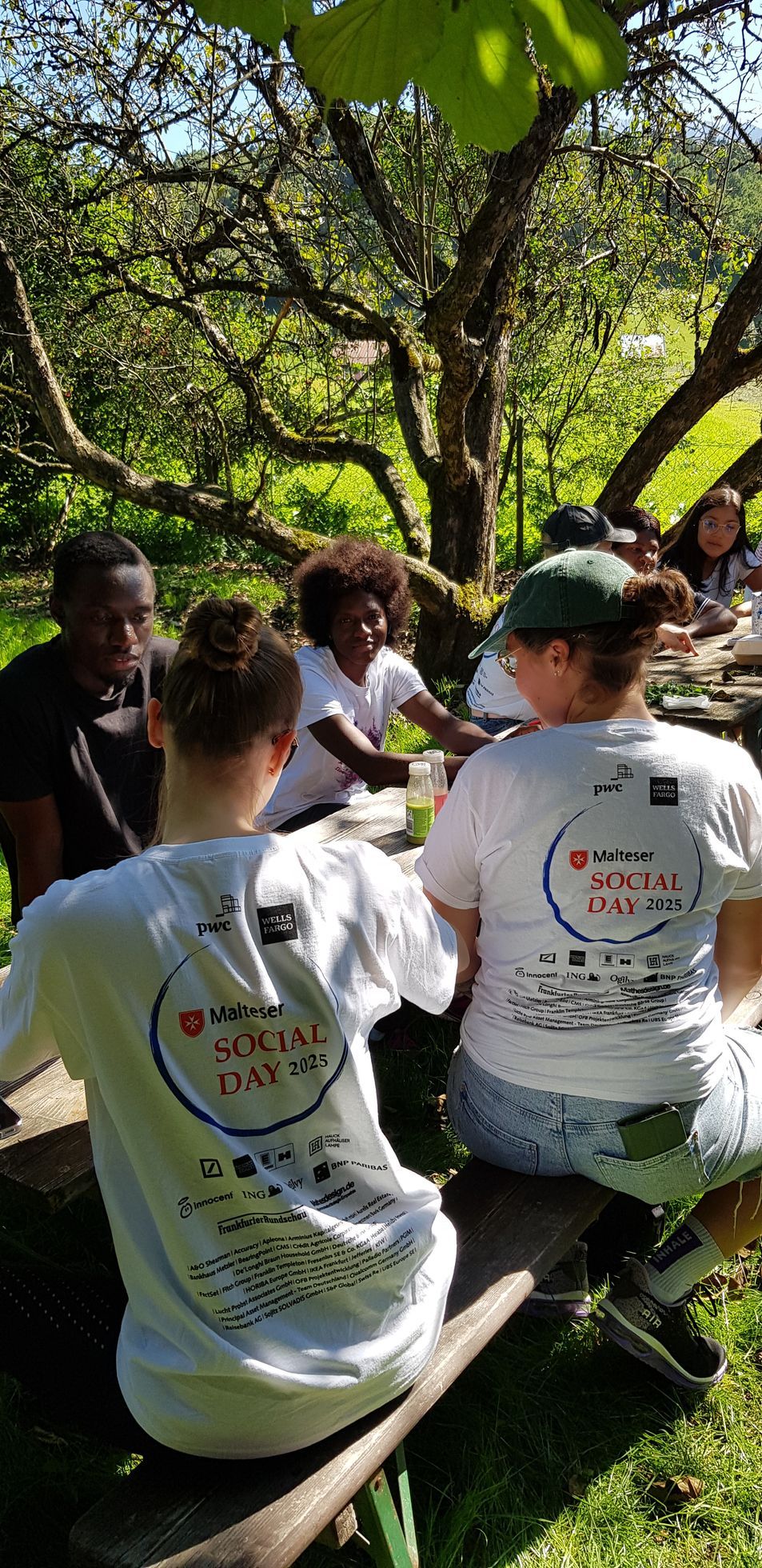 Zwei Personen sitzen auf einer Bank im Freien, tragen weiße T-Shirts mit der Aufschrift 'Malteser SOCIAL DAY 2025' und sind von Bäumen umgeben.