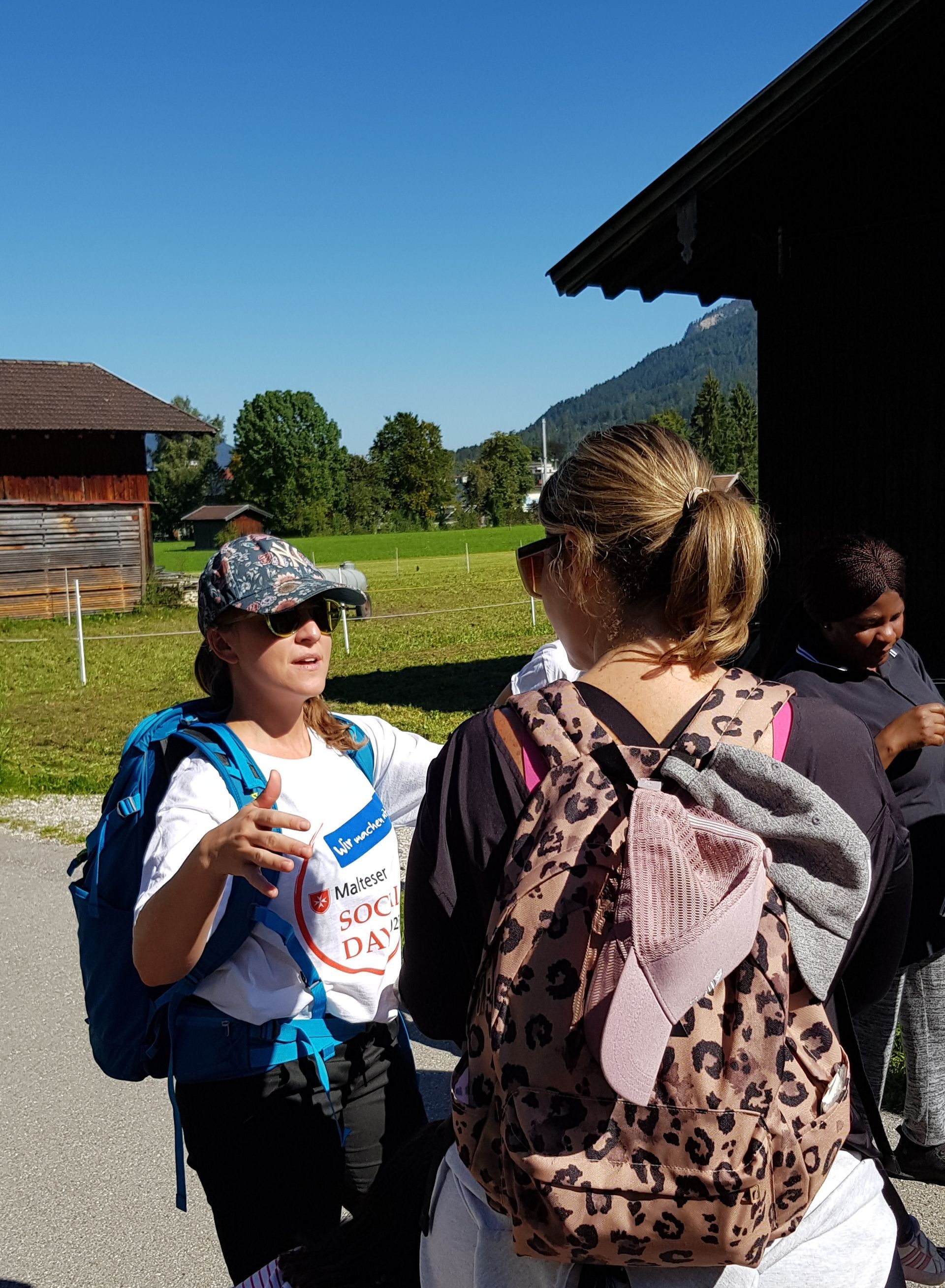 Zwei Frauen im Freien, eine trägt ein weißes T-Shirt mit Aufschrift 'Social Day' und einen blauen Rucksack, die andere einen Leopardenmuster-Rucksack mit einem grauen und einem rosa Kleidungsstück daran.