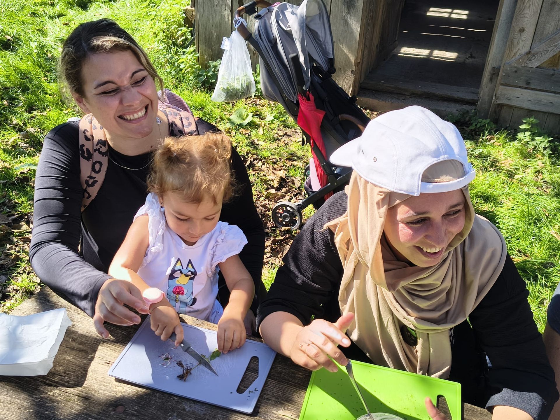 Zwei Frauen und ein kleines Mädchen sitzen an einem Holztisch im Freien, das Mädchen zeigt auf ein Schneidebrett mit Pflanzen, im Hintergrund ein Kinderwagen und eine Holzhütte.