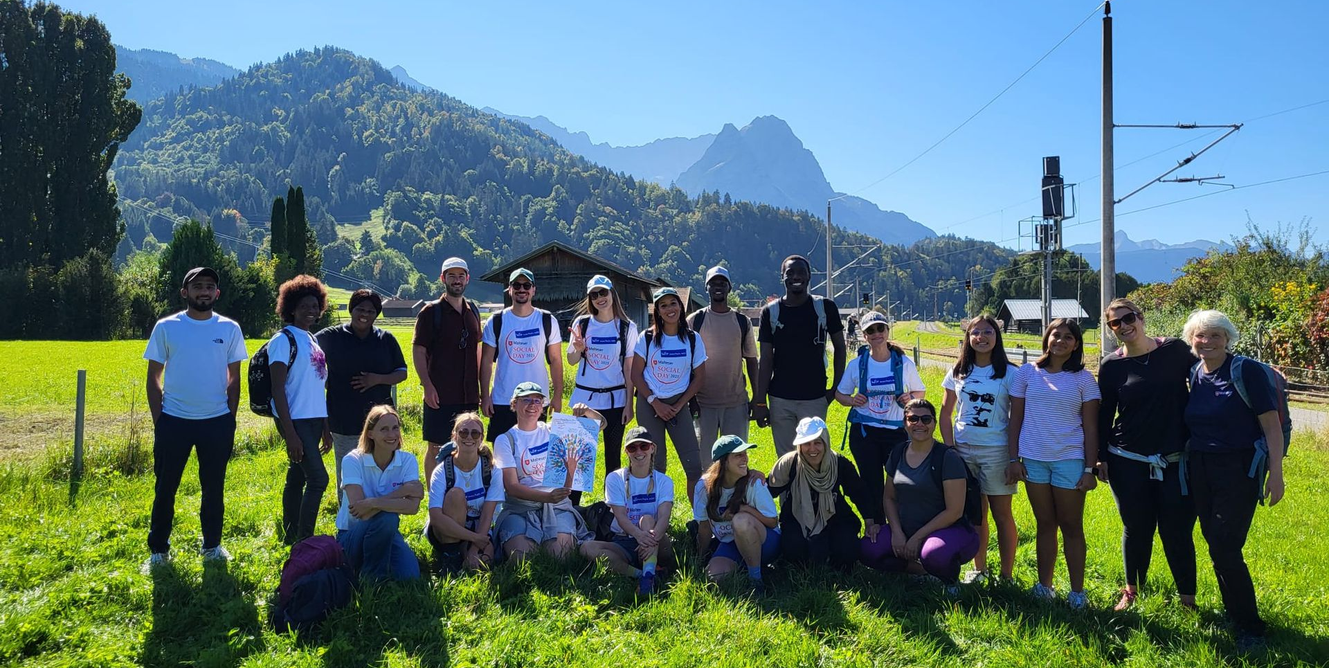 Gruppe von 21 Personen, teils stehend, teils kniend, auf einer grünen Wiese mit Bergen und blauem Himmel im Hintergrund.